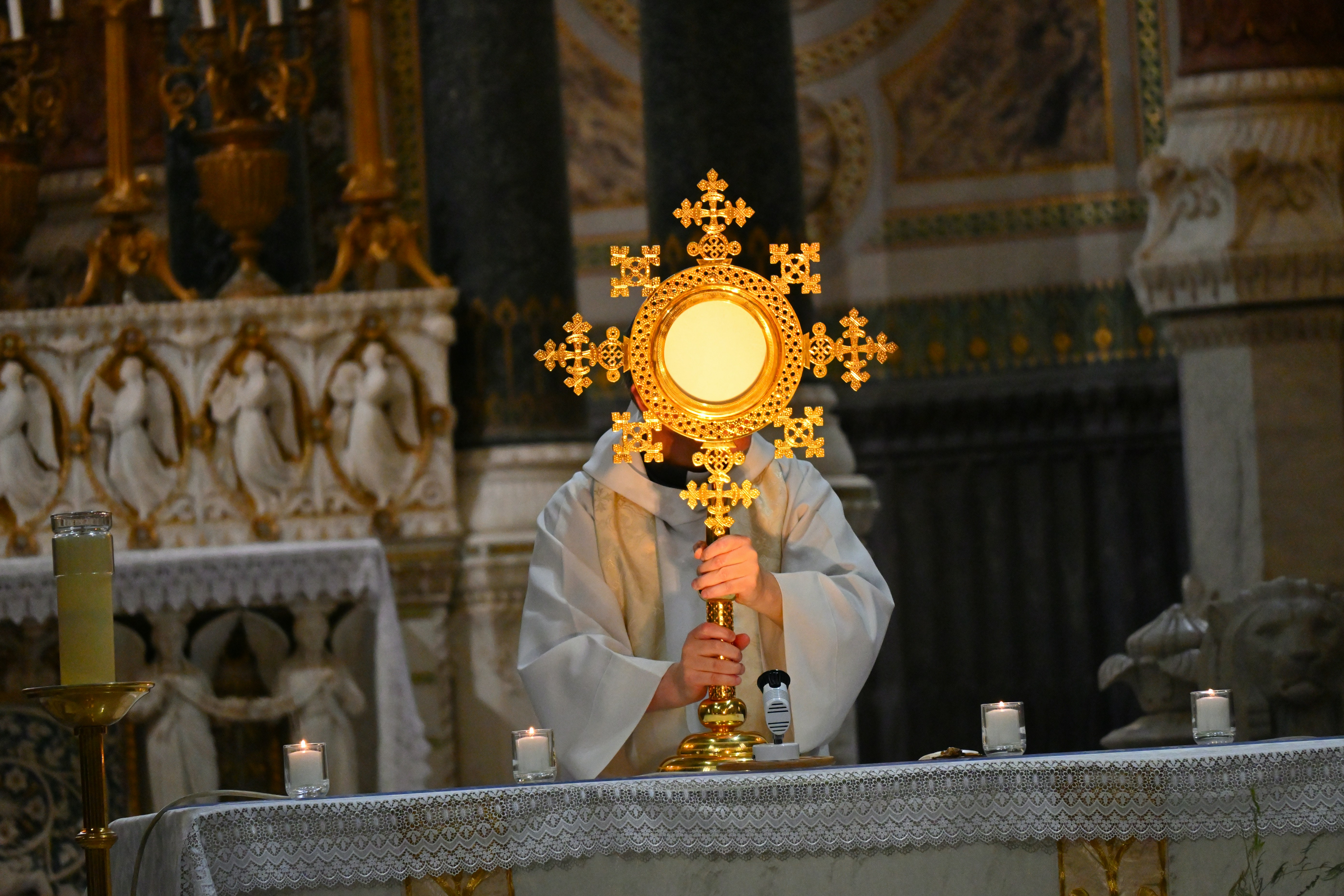 Priest in ornate vestments holding a golden starburst monstrance at an altar during a religious ceremony.