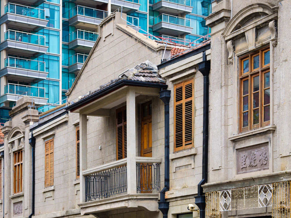 The image shows a two-story building with a gray facade and blue accents, including balconies on both floors. The ground floor features a balcony with wooden shutters, while the upper floor has a balcony with black railings. A sign is visible in front of the building, but its contents are not discernible from the image.