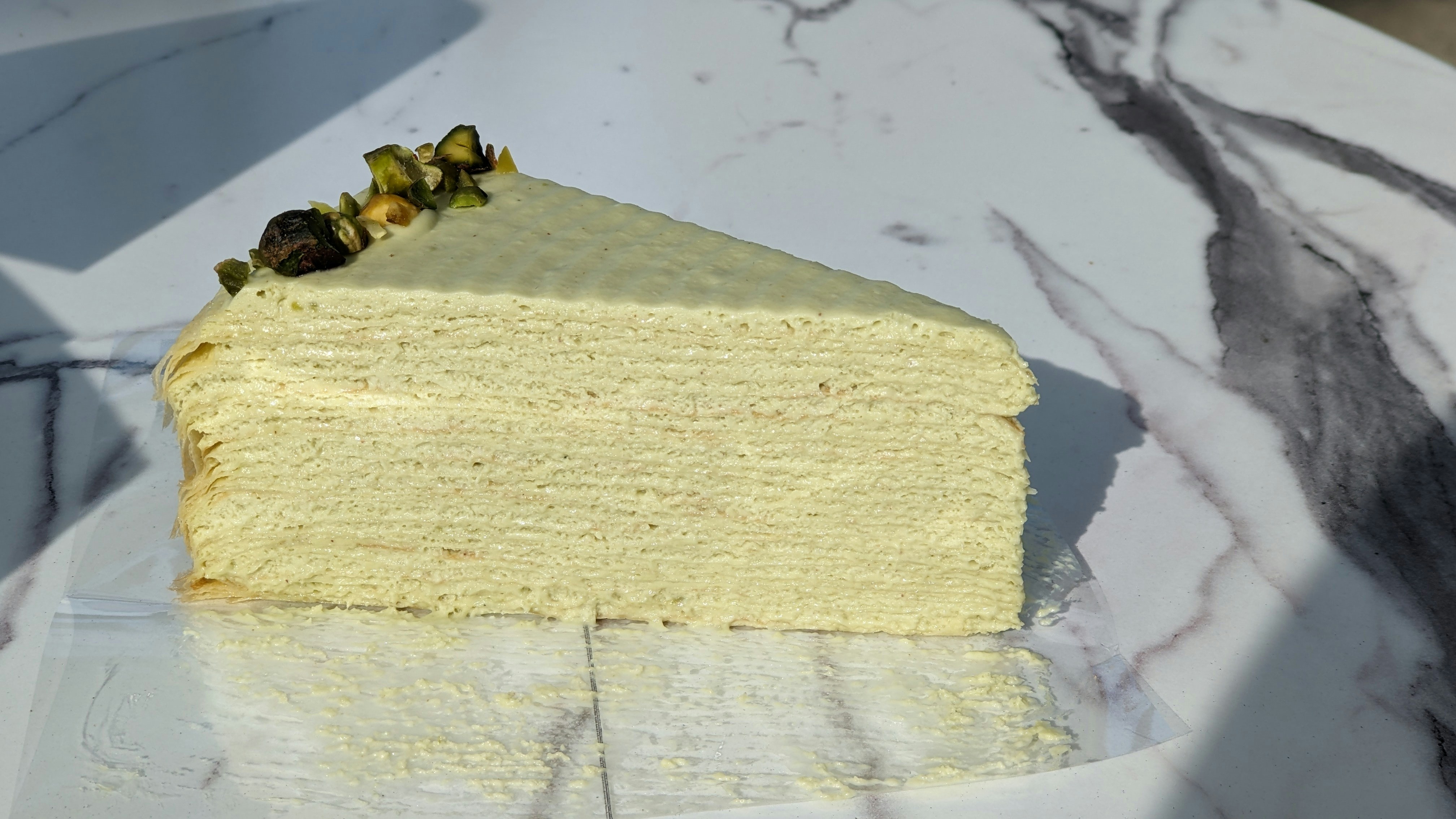 Pistachio cream cake slice with green layers and white frosting on marble surface