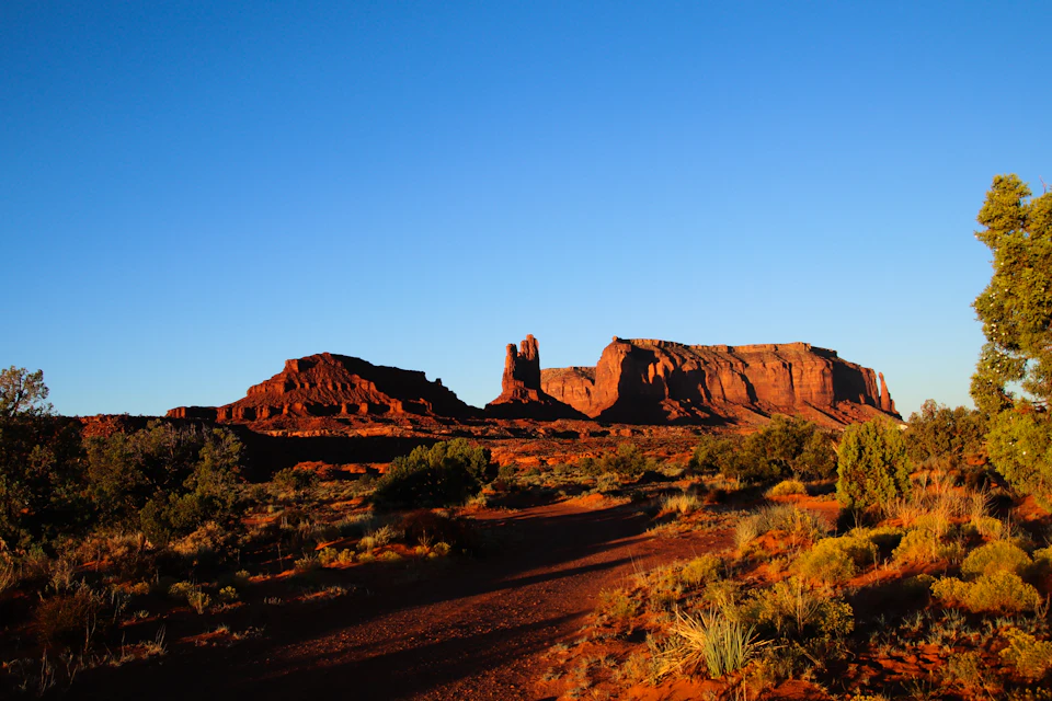 The image depicts a desert landscape with a large, towering red rock formation in the center. The rock formation is surrounded by shrubs and trees, creating a sense of depth and scale. The sky above is a clear blue, providing a striking contrast to the earthy tones below. The perspective of the photo suggests it was taken from a lower angle, looking up at the imposing rock formation.