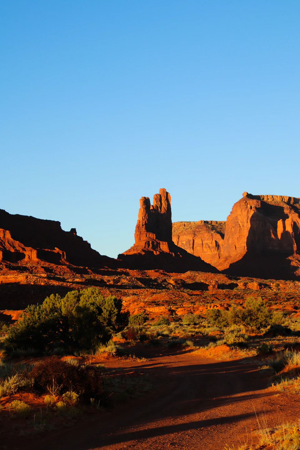The image depicts a desert landscape with a dirt road winding through it, leading towards a tall, rocky formation on the right side. The formation is composed of large rocks and stands out against the clear blue sky. On the left side of the road, there are trees scattered across the scene, adding a touch of green to the otherwise arid environment.