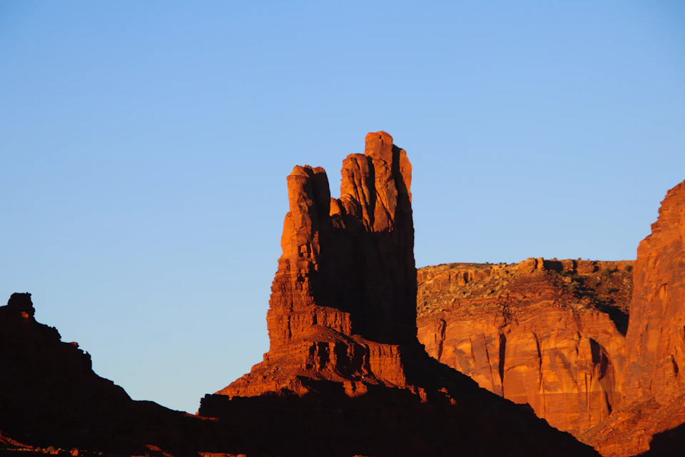 The image depicts a majestic rock formation with three distinct layers, each exhibiting unique colors and textures. The largest layer is a reddish-brown hue, while the middle layer has a lighter shade of brown, and the smallest layer is a darker red color. These rock formations are situated in a desert landscape, with a clear blue sky providing a striking backdrop to their rugged beauty.

The image is taken from a low angle, emphasizing the towering height of the rock formation as it reaches towards the sky. The sun's rays shine down on these rocks, casting long shadows and highlighting their intricate details.