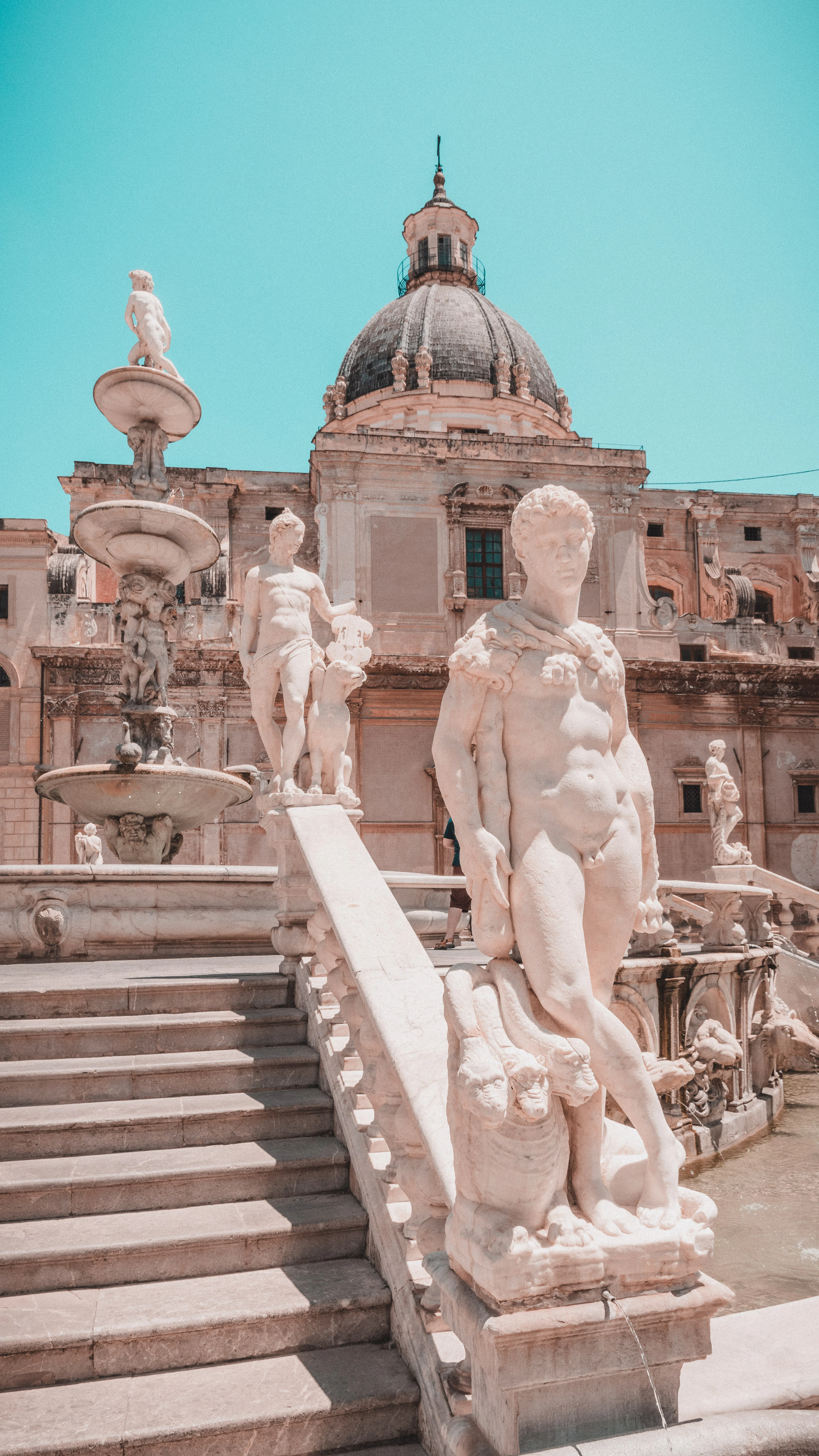 White marble nudes and tiered basins of the Fontana Pretoria in Palermo, with the grey stone dome of Santa Caterina rising behind them under a clear turquoise sky.