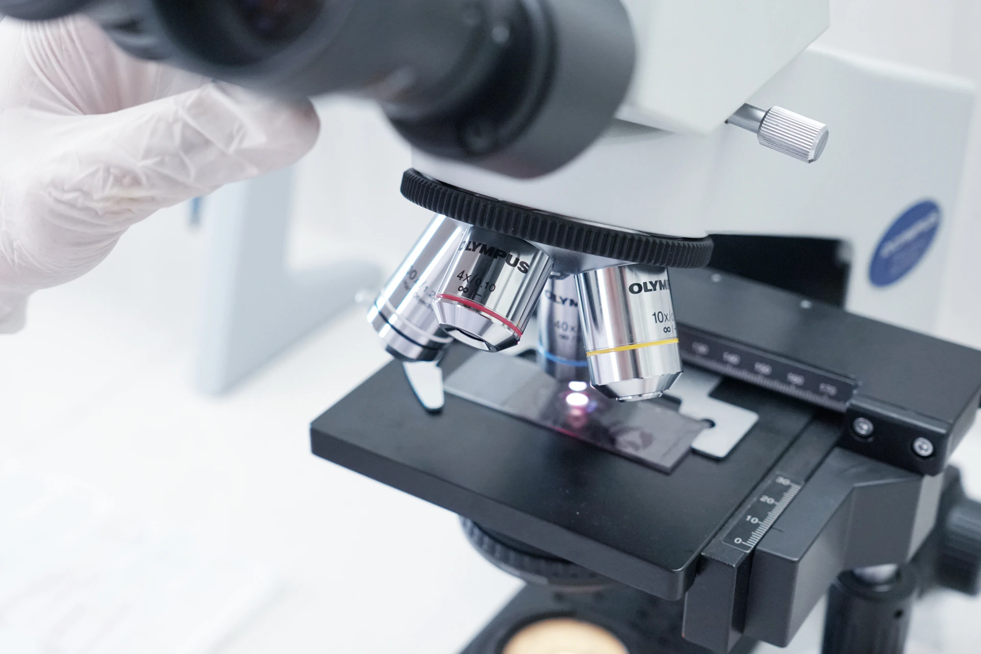 A person in white gloves is looking through a microscope