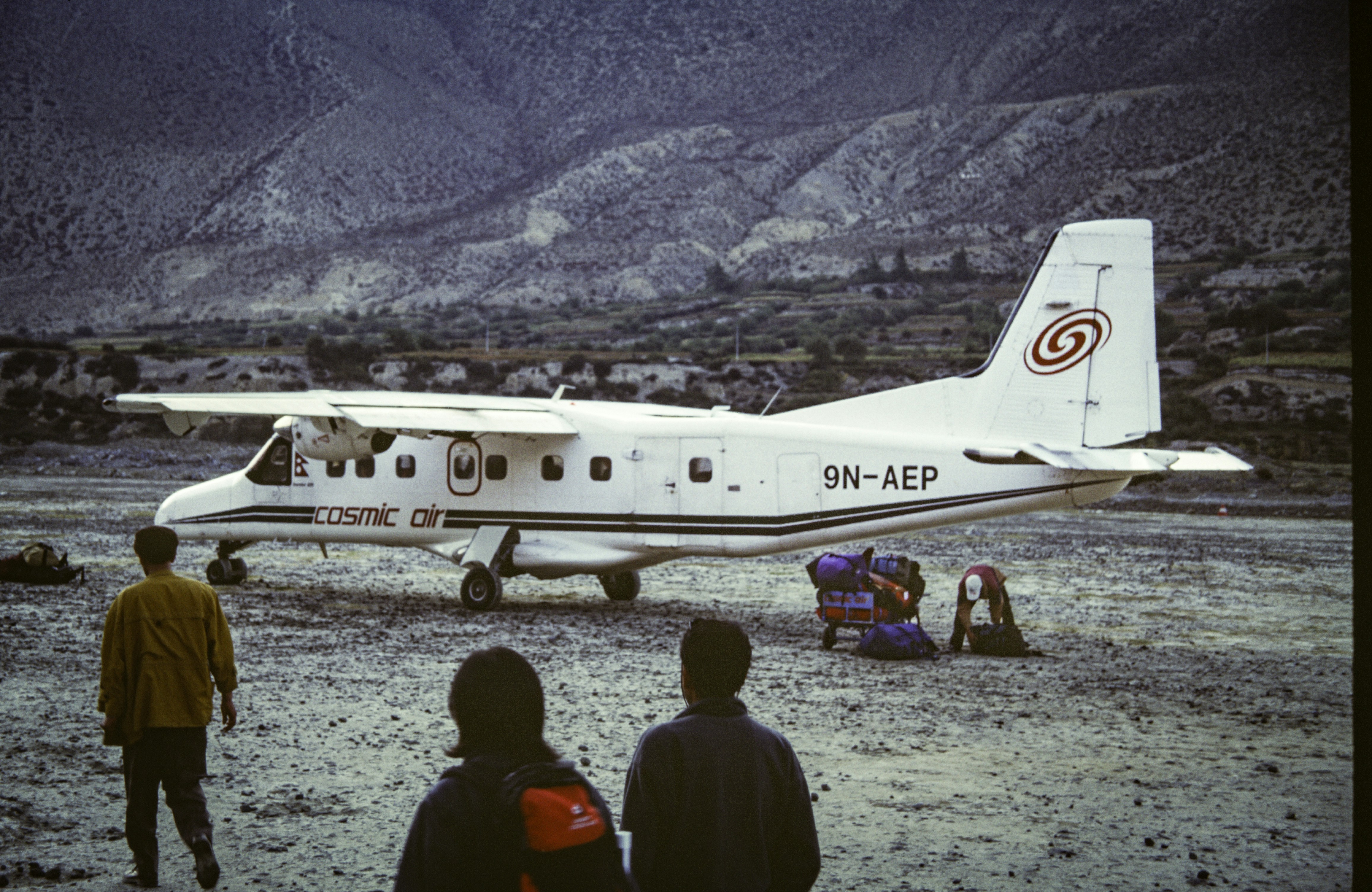 A group of people standing around a small plane, A small twin-engine airplane from Cosmic Air is parked on a rough, gravel landing strip surrounded by a mountainous landscape. A few passengers stand nearby, with backpacks and cargo loaded next to the plane. The plane’s registration number, 9N-AEP, is visible on its side. The scene depicts a remote rural airport, possibly in the Nepalese highlands, with minimal infrastructure and rugged terrain in the background.