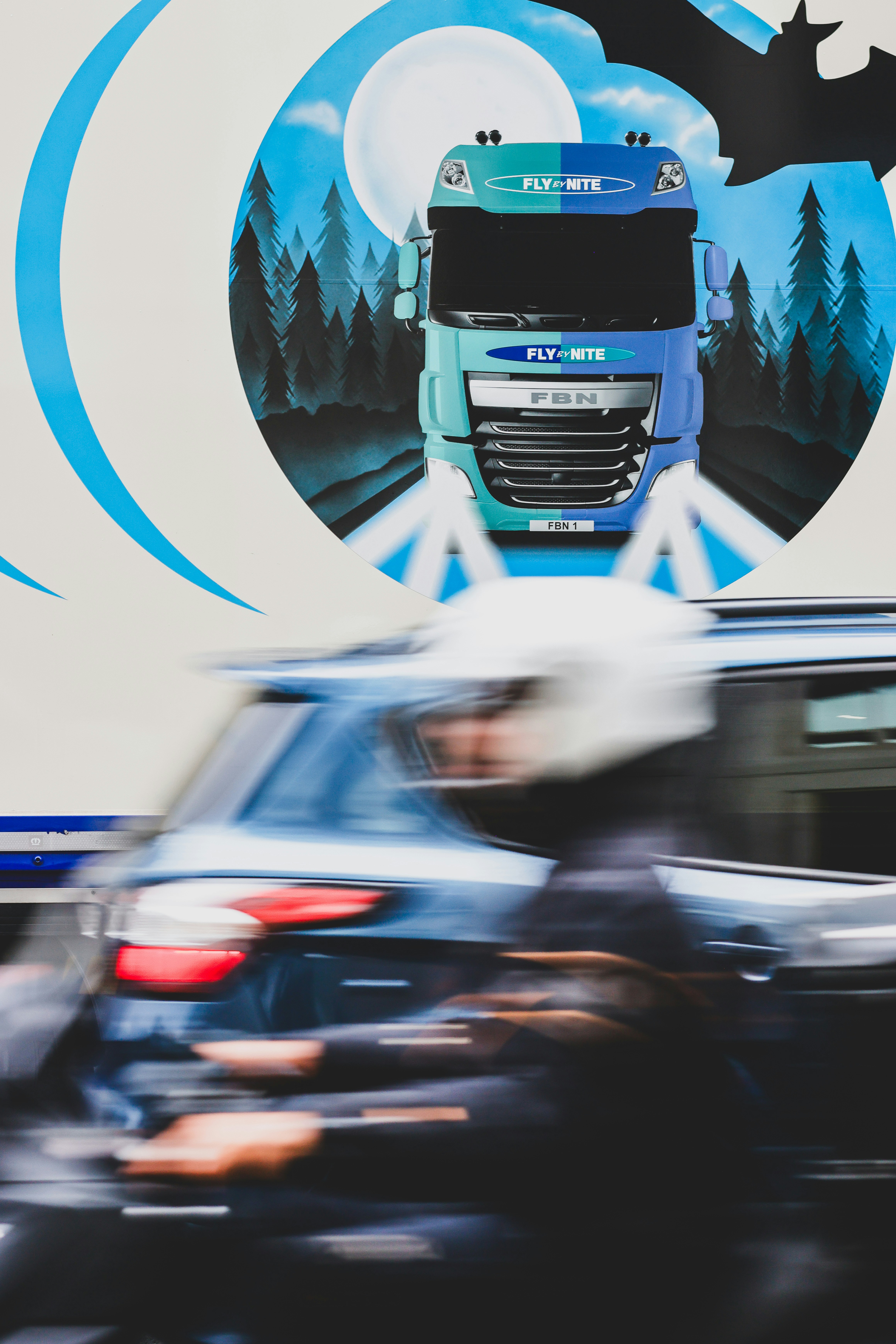 Photograph featuring a circular blue logo backdrop with a branded truck in the background. Foreground vehicles are blurred by motion, emphasizing the dynamic scene.