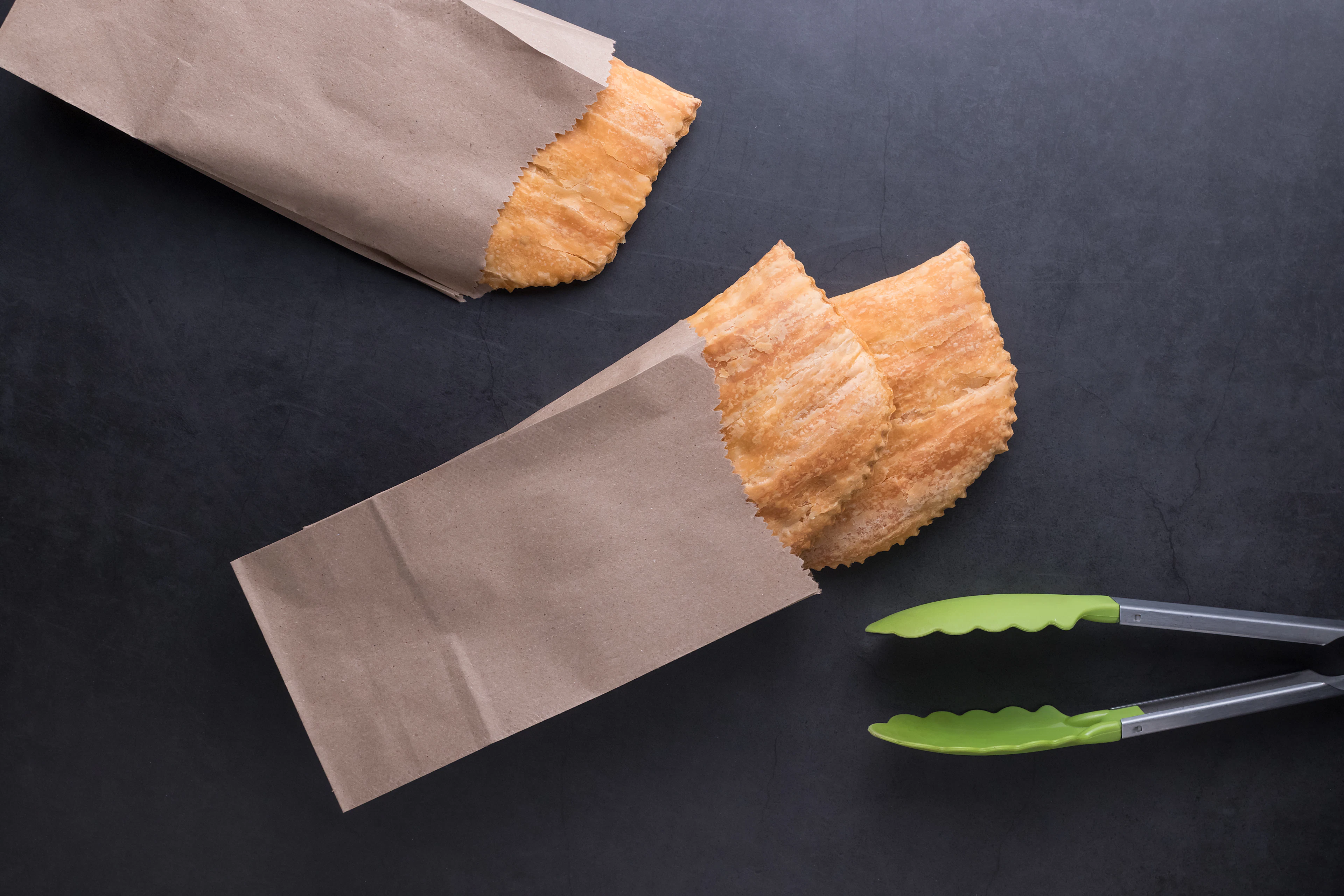 Pastries in small paper bags on a dark bench with serving tongs