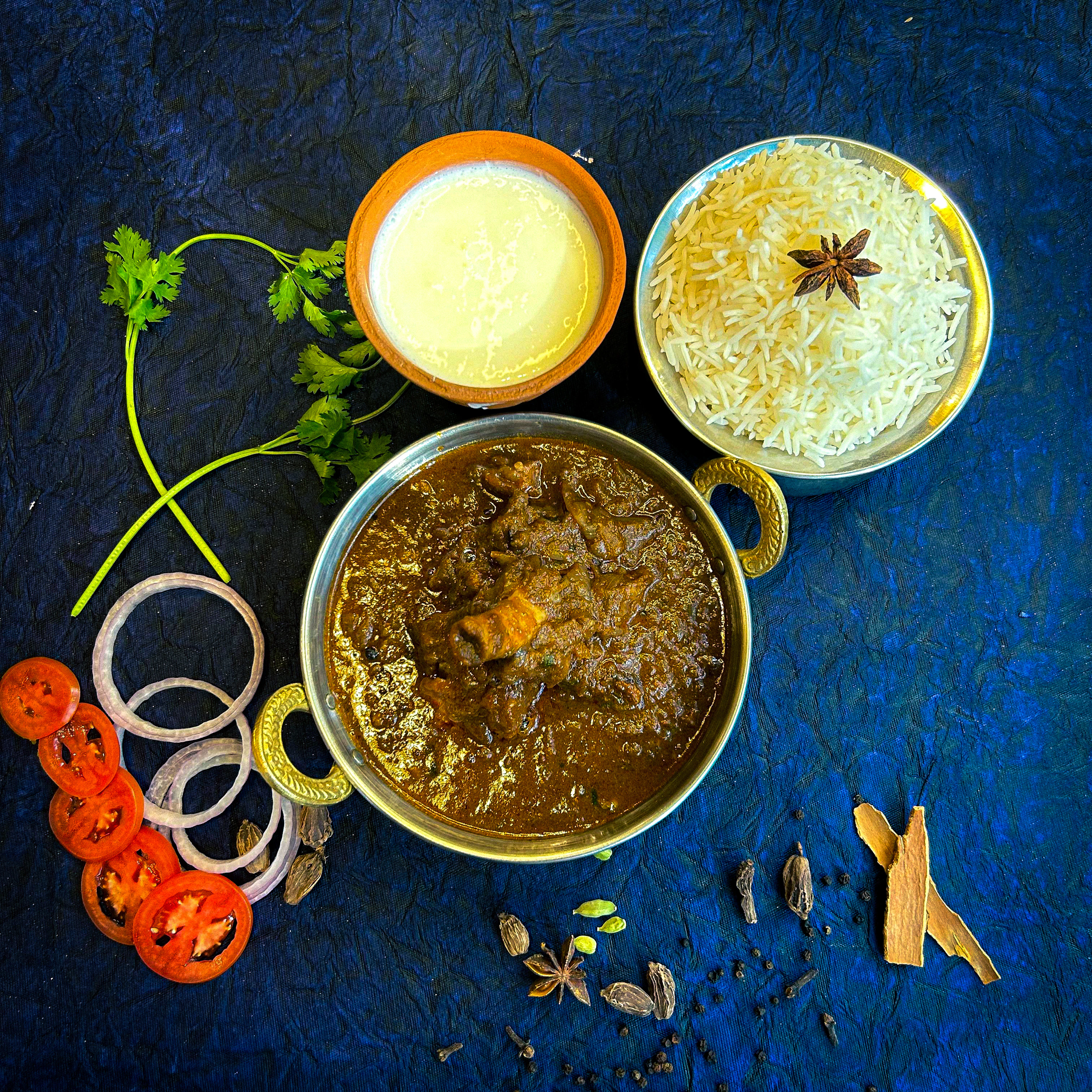 A dark North Indian mutton curry in a brass kadai, served with star-anise basmati rice, a clay cup of lassi, sliced onion and tomato, whole spices scattered alongside