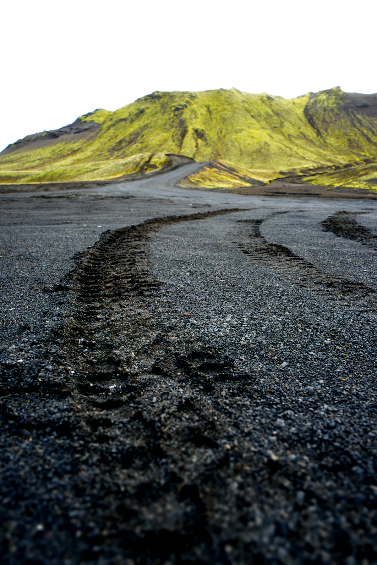 Tire tracks in black gravel on a highland road, curving toward a moss-green hill