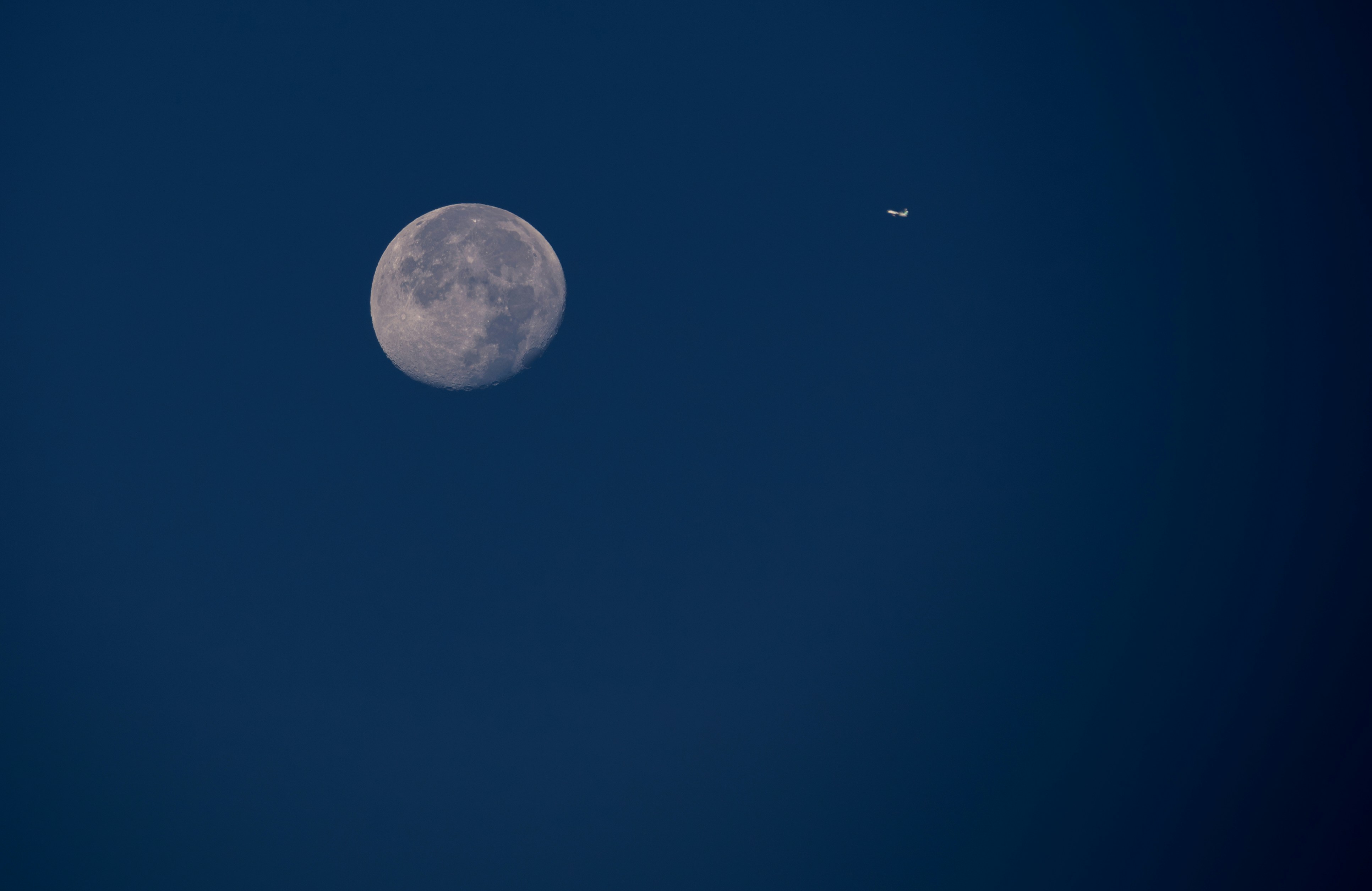 A full moon and a plane flying in the sky, A nearly full moon dominates the dark blue sky, with an airplane visible flying in the distance. The moon