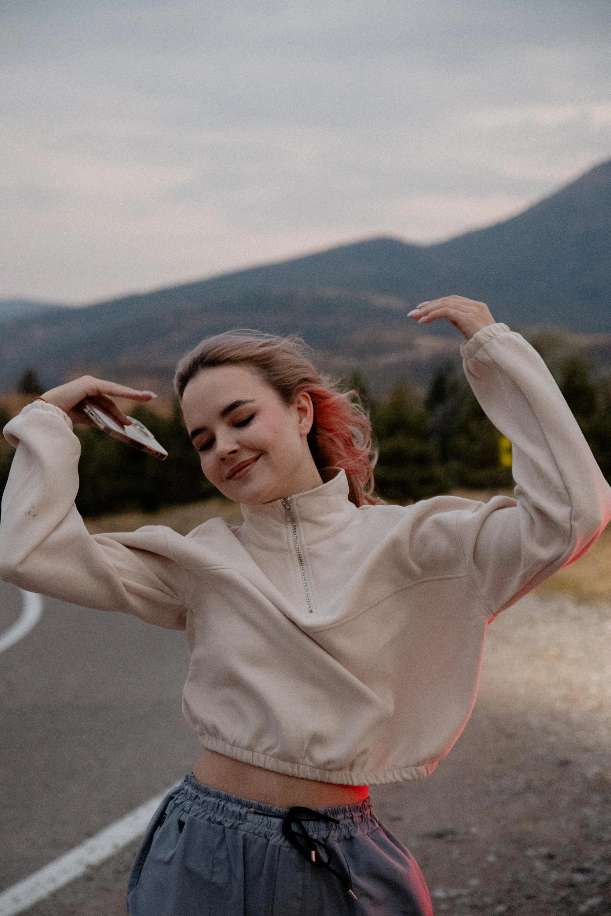 A woman in a cream sweater dancing on a mountain road
