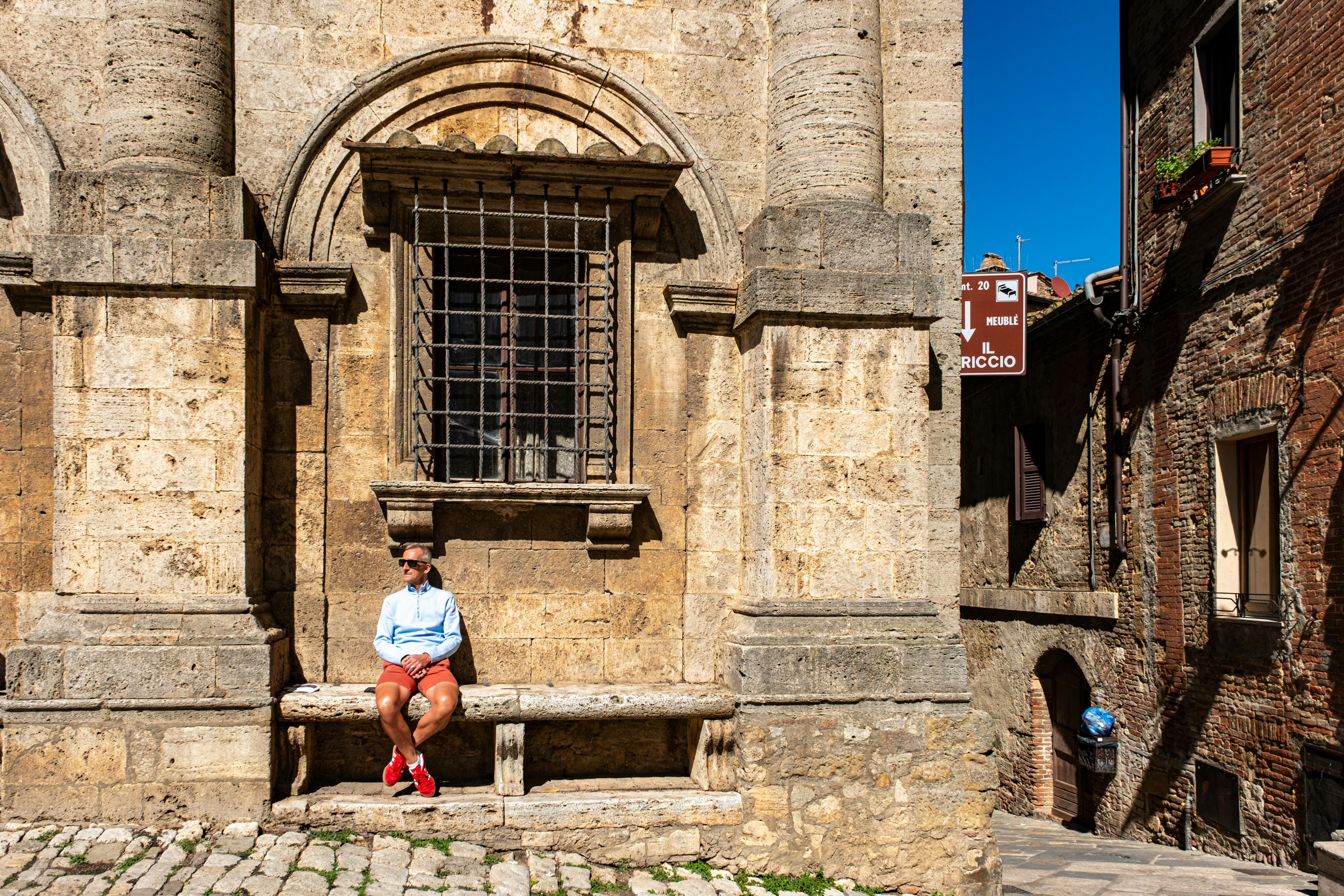 A person sitting on a bench in front of a building, A tourist waits on a stone bench in front of one of the ancient buildings in the main piazza of the medieval and Renaissance town of Montepulciano in Tuscany, Italy. Many of the buildings that give the Piazza Grande its distinctive character were constructed between the 14th and 16th centuries. (model release applies)