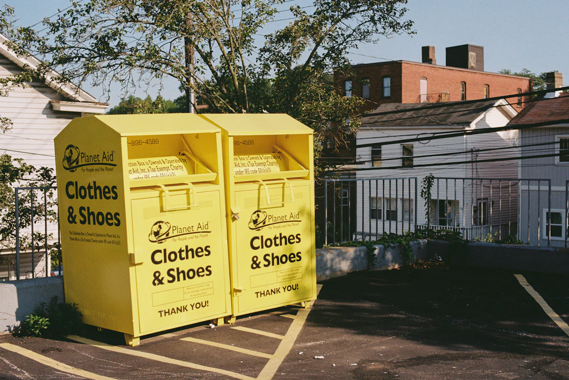 Two yellow steel clothing collection banks standing together at the edge of a car park.