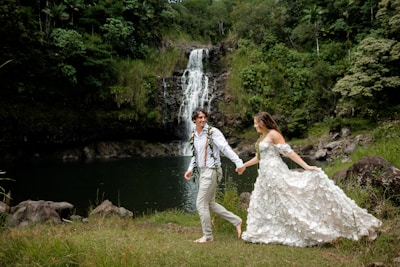 A man and a woman in a wedding dress holding hands near a waterfall