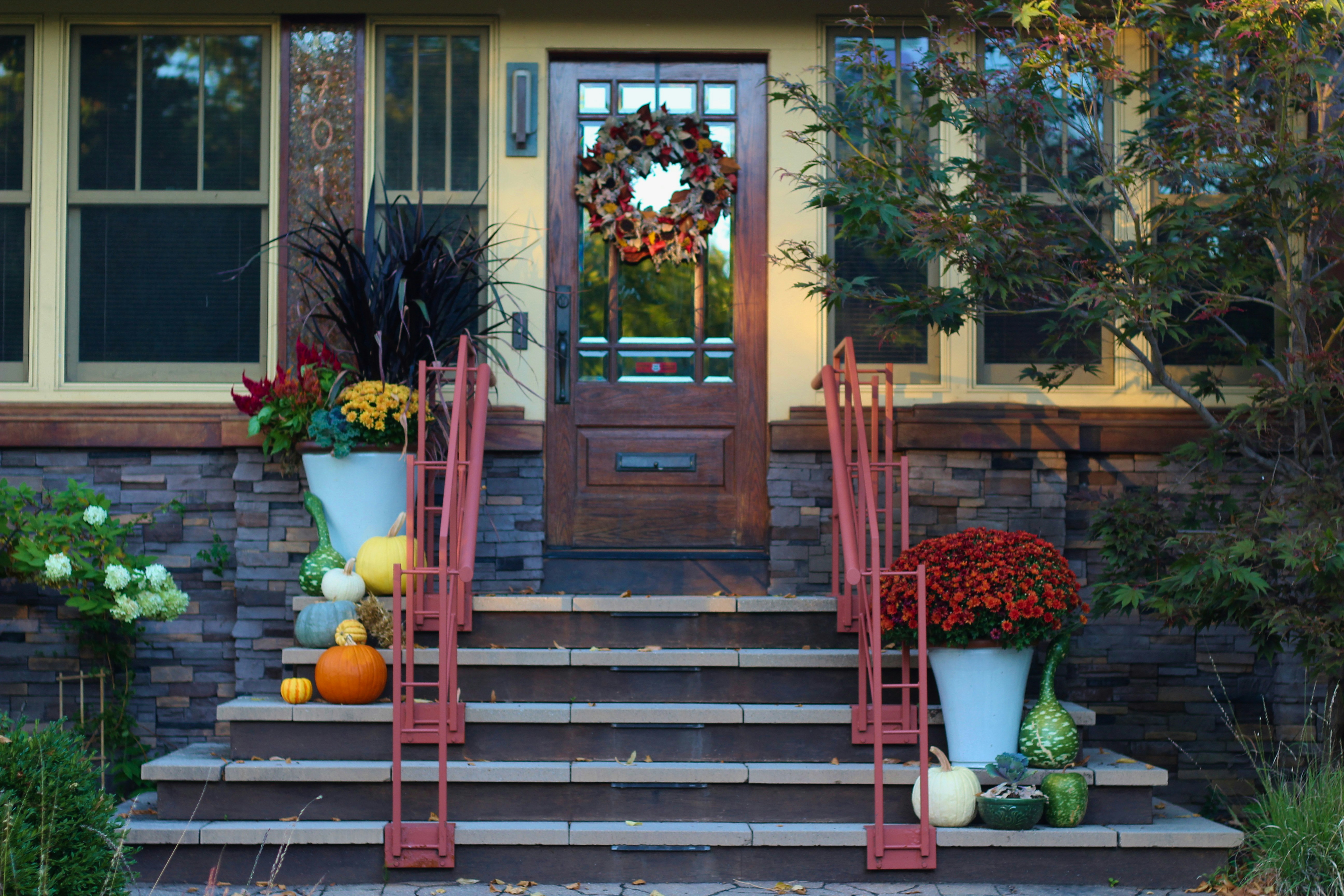 A charming front porch decorated for autumn. The front door is adorned with a fall wreath and flanked by potted mums. The steps are lined with planters filled with pumpkins, gourds, and flowers. The house has a stone facade and large windows.