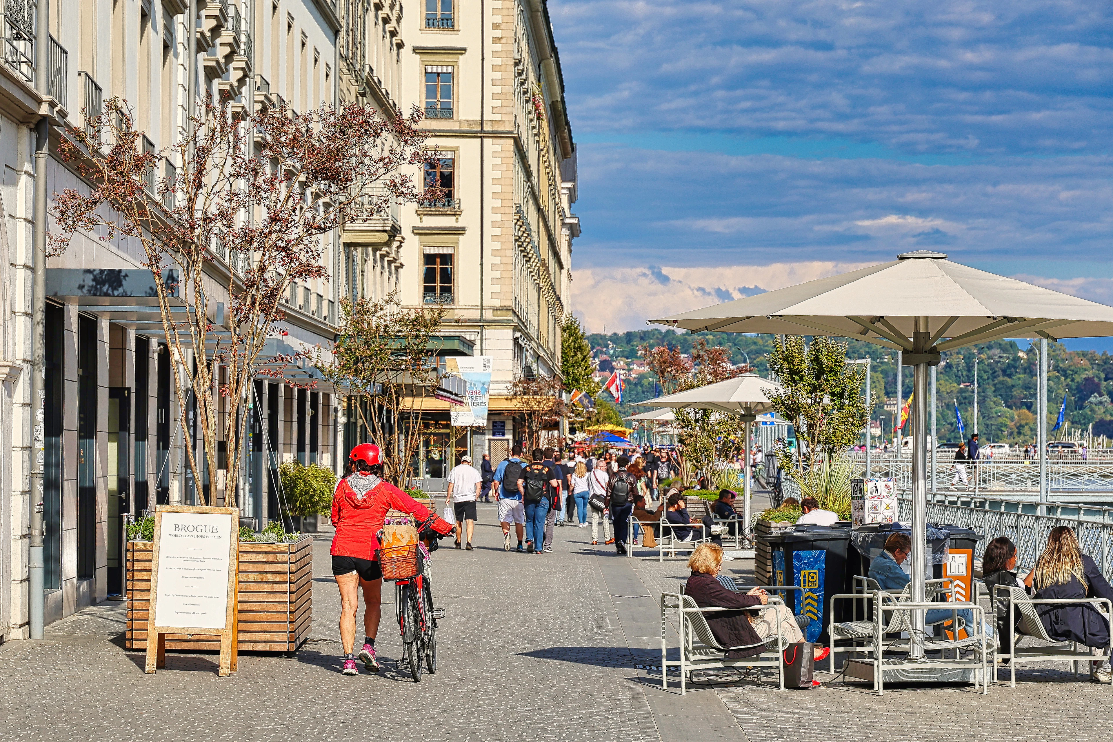 The Quai des Bergues pedestrian promenade in Geneva on a sunny day: a cyclist passing café terraces with white parasols and loungers, people walking beside the water