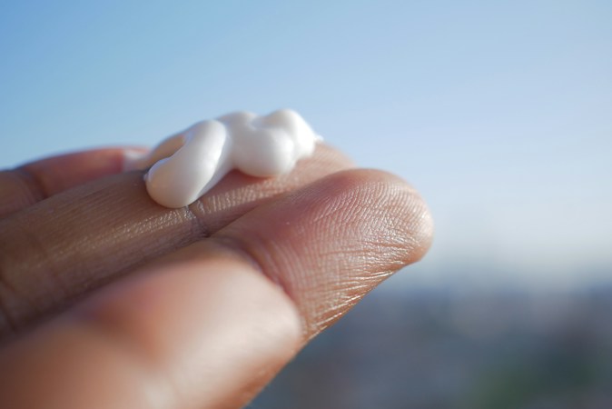 Close-up of a fingertip holding a dollop of white sunscreen against a pale blue sky