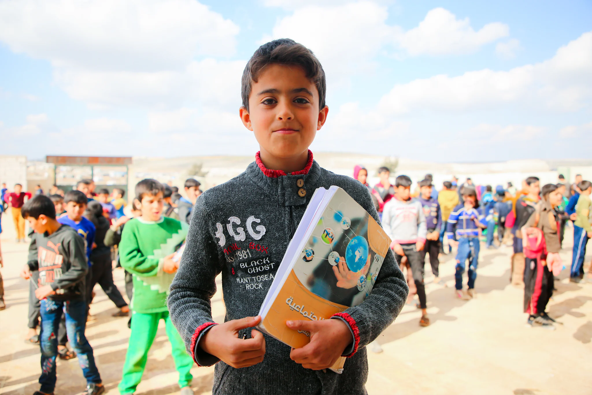A boy holds his school textbooks in a camp schoolyard