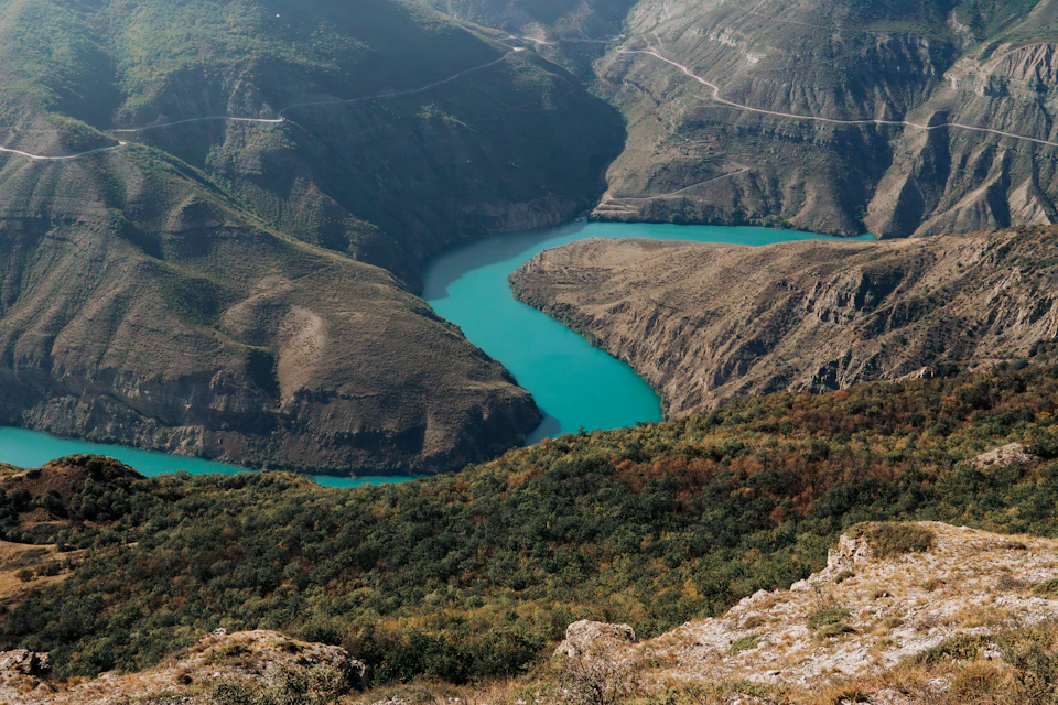 The image depicts a breathtaking view of a deep blue river winding through a mountainous landscape. The river, with its vibrant turquoise color, flows gracefully from left to right across the frame. The mountains, painted in various shades of brown and green, rise majestically on either side of the river, creating a sense of depth and scale.

The sky above is a light blue canvas dotted with fluffy white clouds, adding to the overall serene atmosphere of the scene. The image is taken from an elevated perspective, providing a bird's eye view of this picturesque landscape.