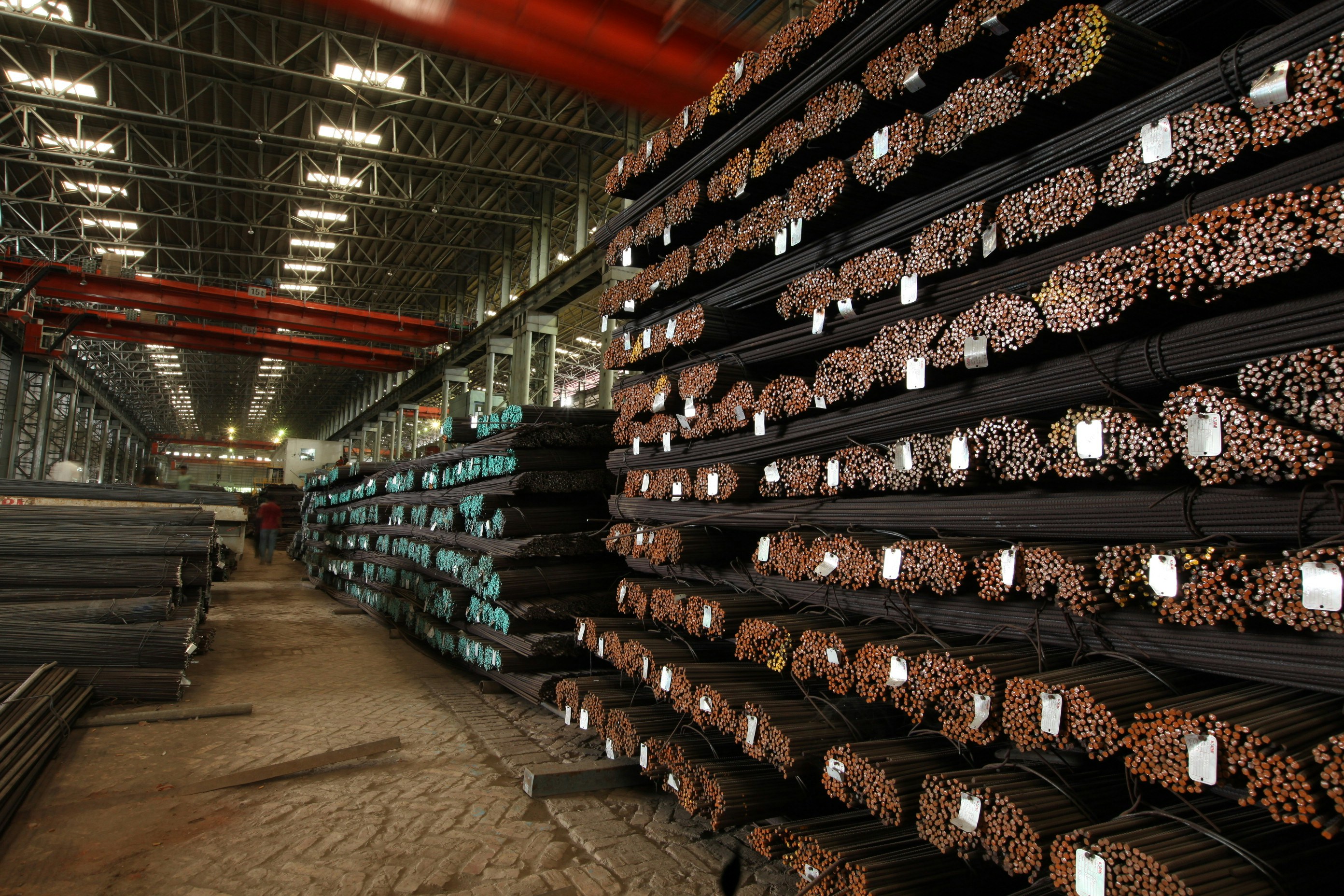Large industrial warehouse interior with steel inventory stacked in rows, bright overhead lights illuminating dark steel sections