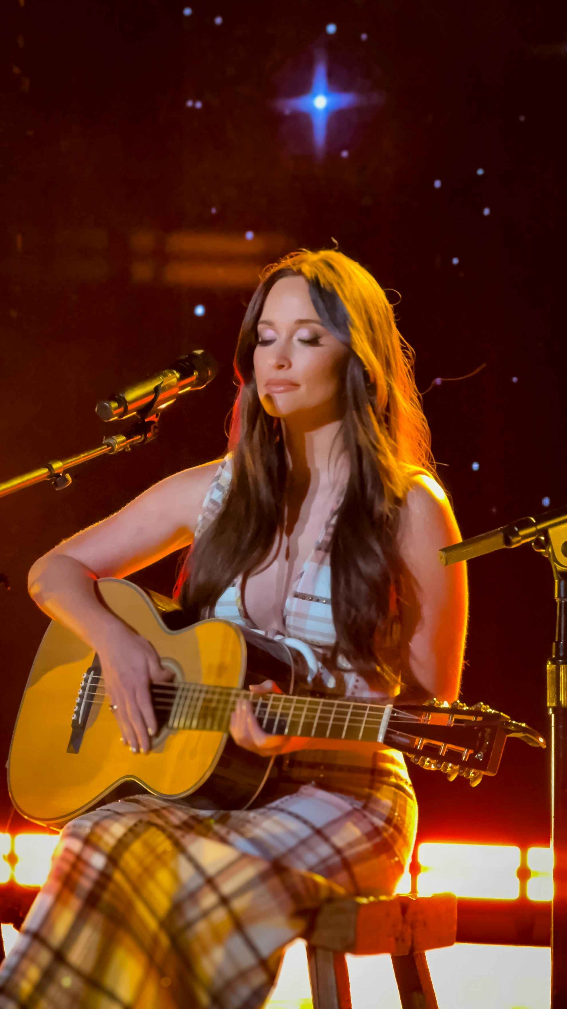 Female singer with acoustic guitar on stage, warm stage lighting, rustic wooden backdrop