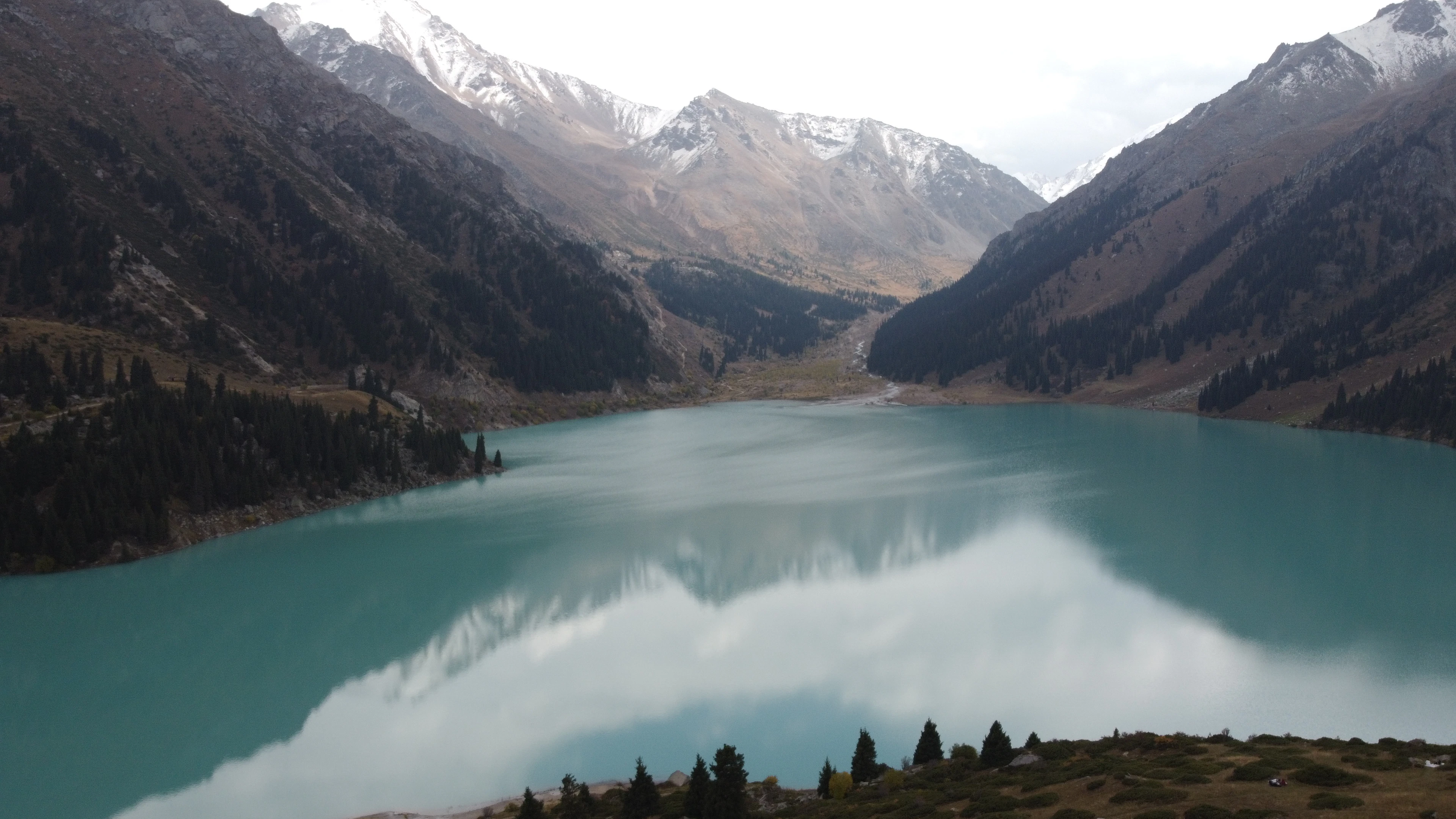 Big Almaty Lake in the mountains above Almaty, its milky turquoise water held between steep spruce-covered slopes with the first snow lying on the peaks above