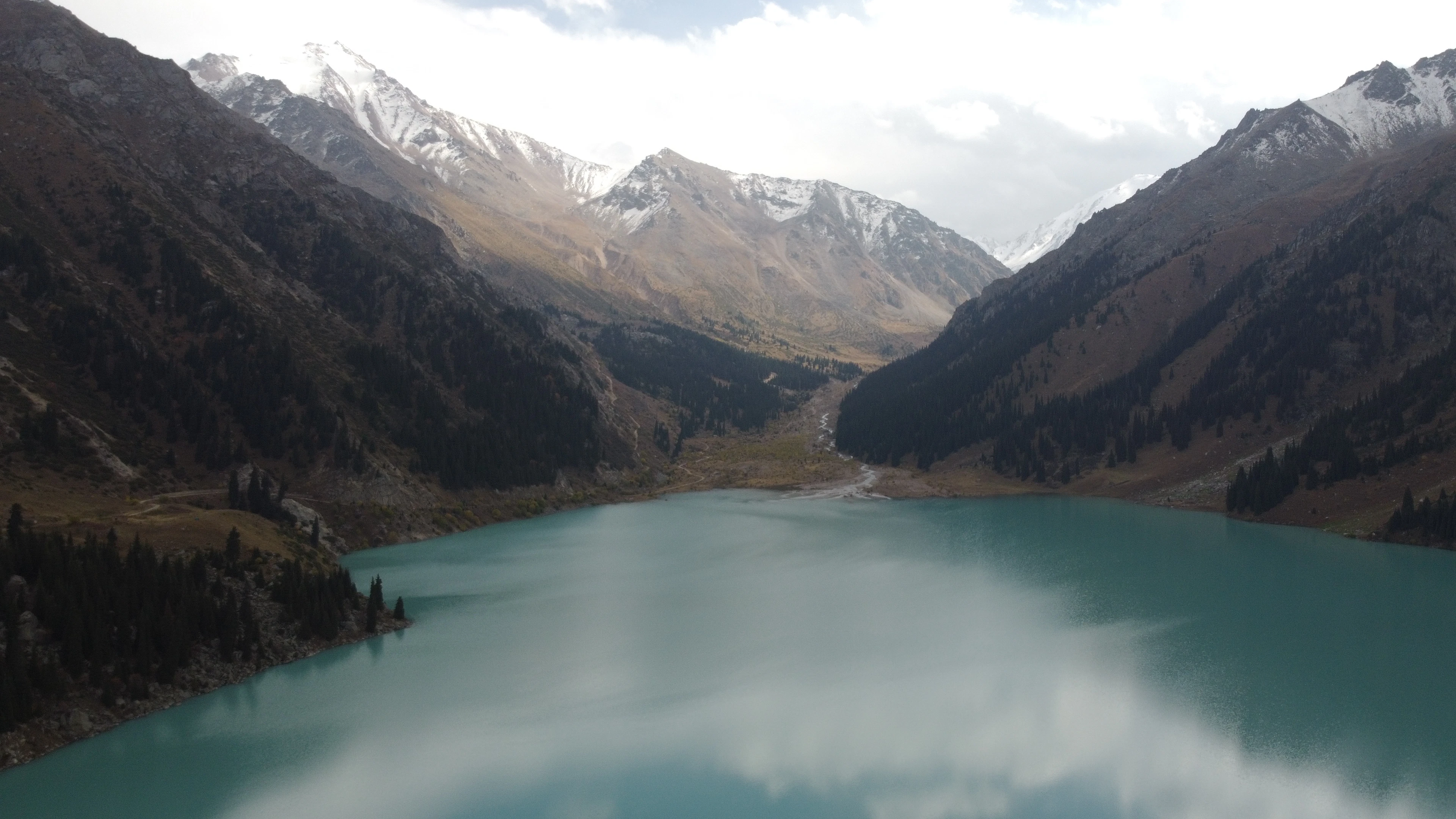 The turquoise waters of Big Almaty Lake framed by steep, snow-dusted mountains under a cloudy sky.