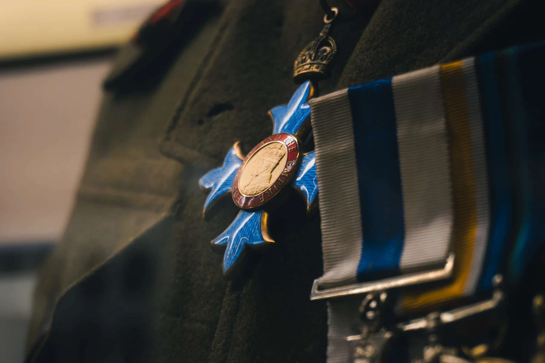A blue enamel cross with a gilt center hanging on a khaki uniform beside striped ribbons