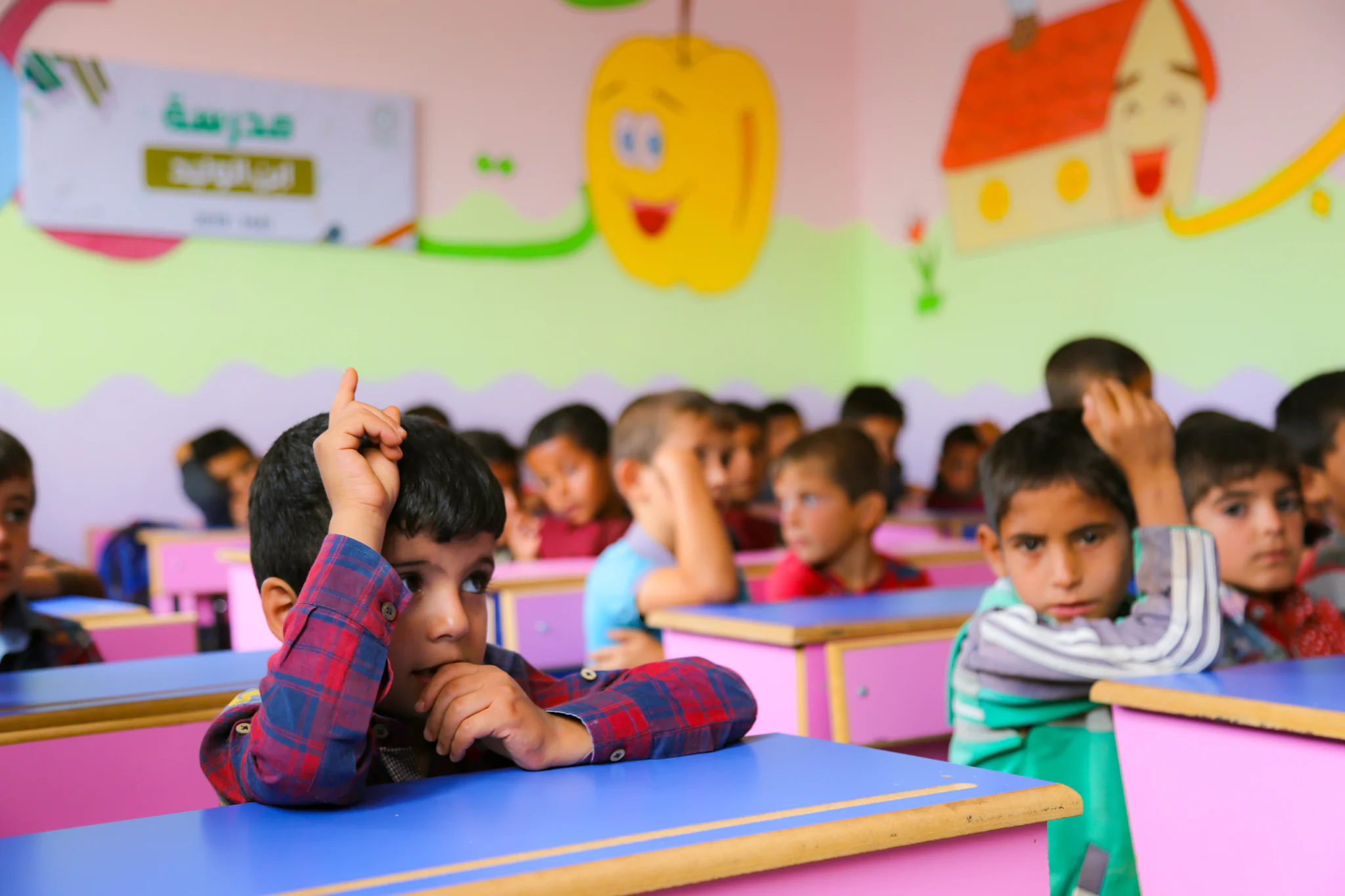 A boy raises his hand in a brightly painted classroom full of pupils