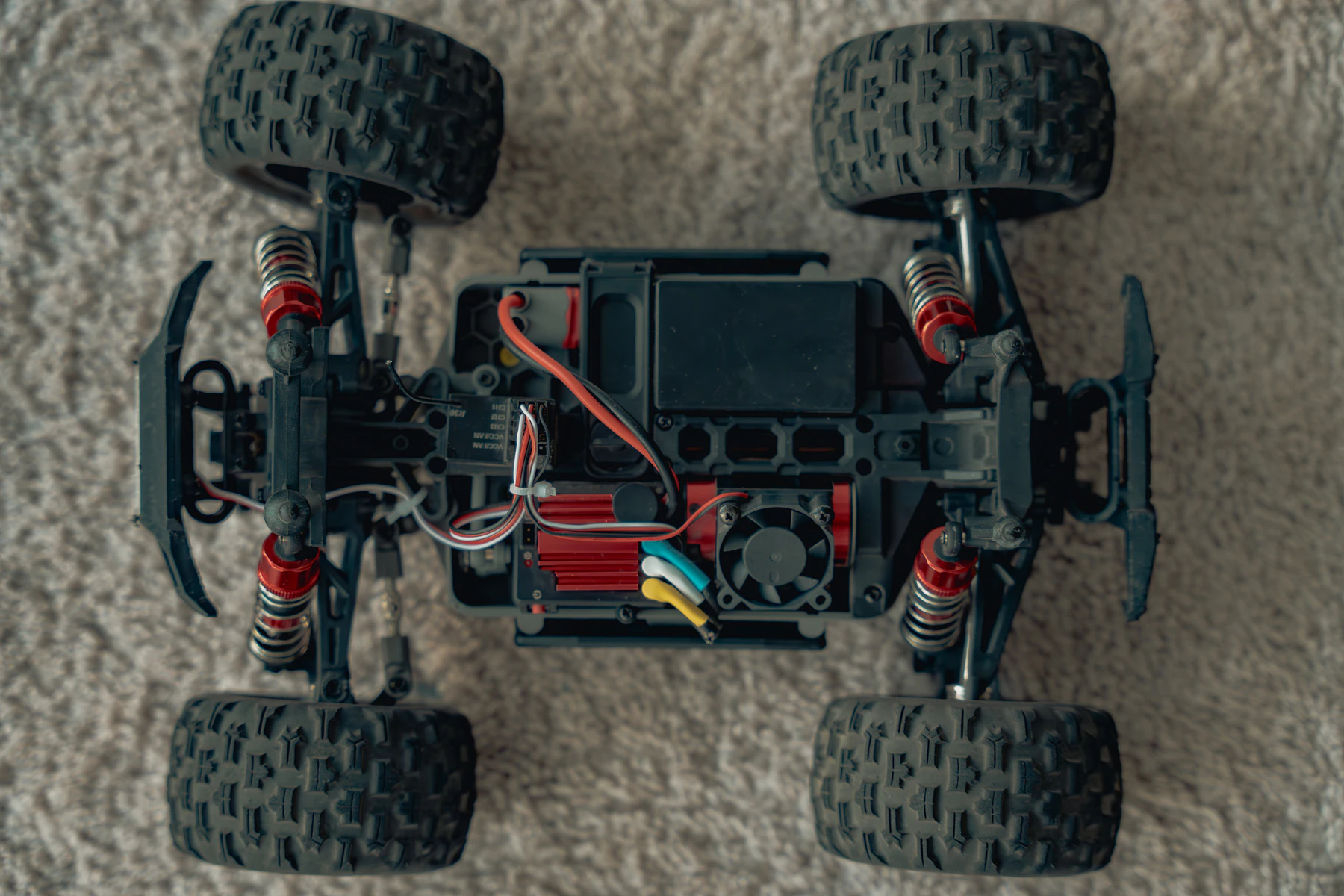 A truck chassis seen from above with the body off, showing four knobby tires, red shocks, wiring and a cooling fan