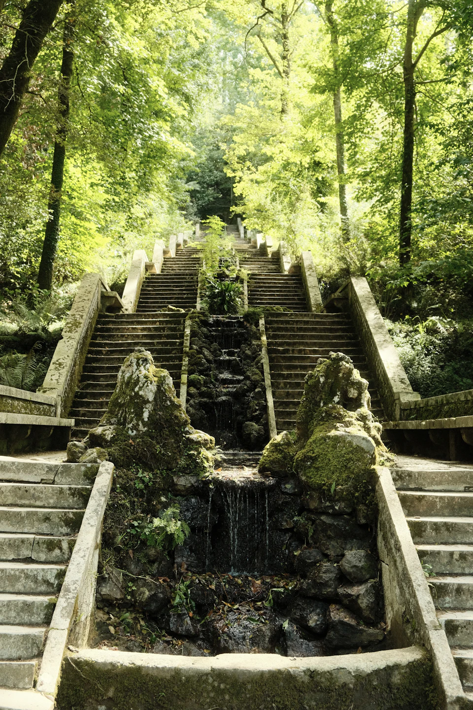 The image depicts a stone staircase leading up to a waterfall, surrounded by lush greenery and trees. The stairs are made of large rocks and appear weathered over time. A small pool of water is located at the base of the stairs, with moss growing on it. The waterfall cascades down from the top of the stairs towards the bottom, creating a sense of depth in the image.