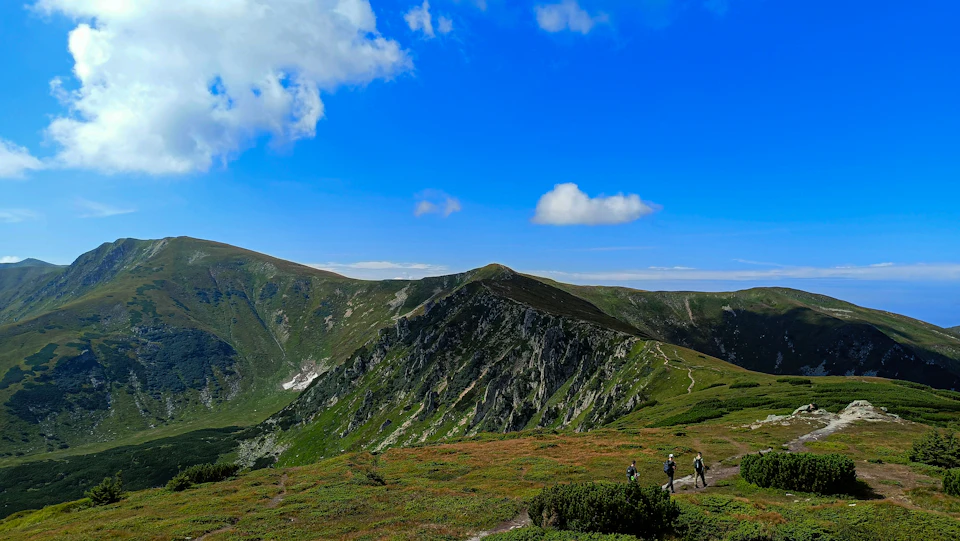 The image depicts a group of hikers ascending a mountain trail surrounded by lush green vegetation and towering mountains under a clear blue sky with fluffy white clouds. The hikers are moving towards the right side of the frame, their backpacks visible on their backs as they navigate the challenging terrain.