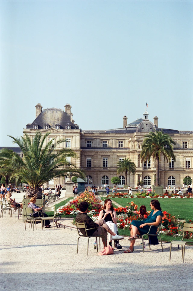 Visitors relaxing in green garden chairs on the lawn in front of the Luxembourg Palace on a sunny day.