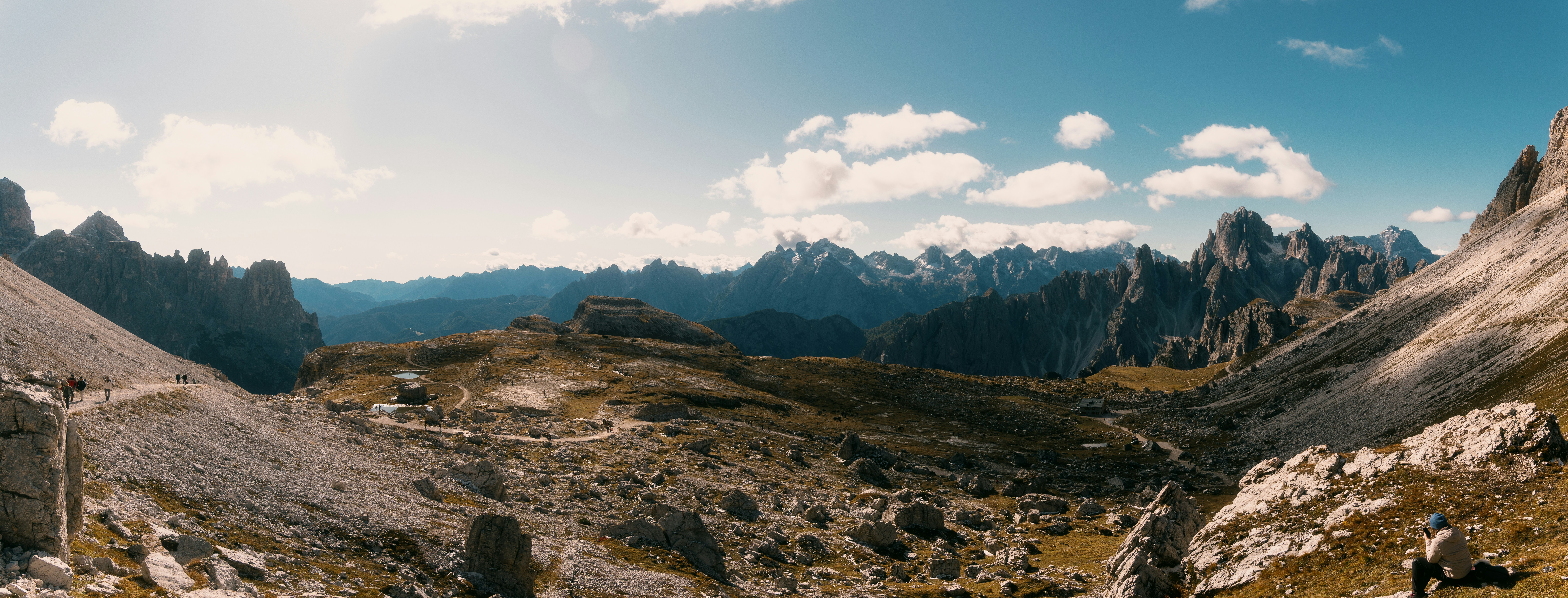 Mountain hiking trail at high altitude with dramatic rocky peaks, crisp blue sky, adventurers in distance