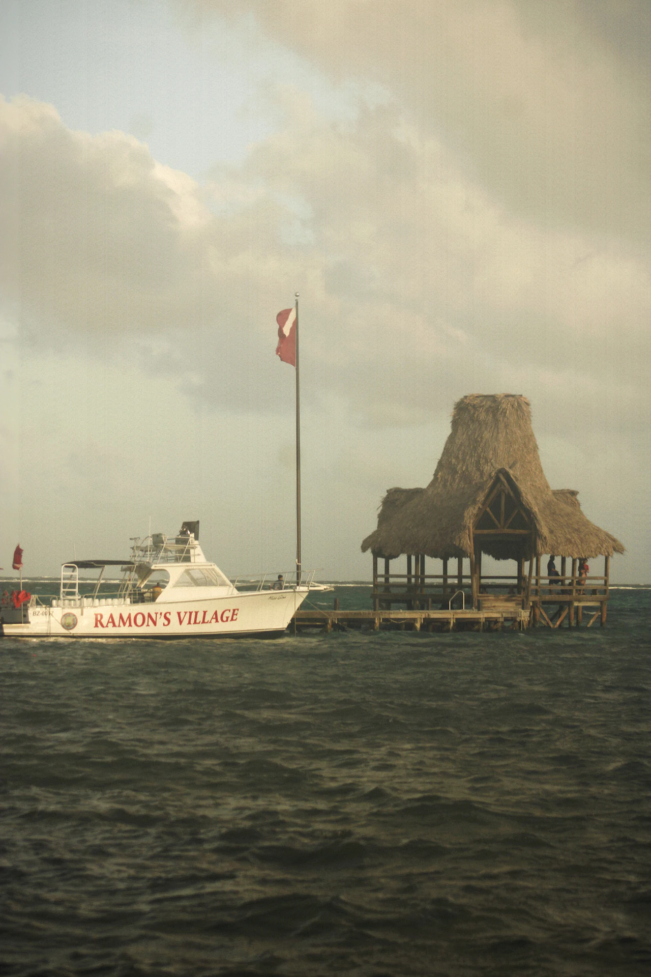 A white boat moored beside a thatched-roof pier in grey-green water under an overcast sky, with a red flag flying.