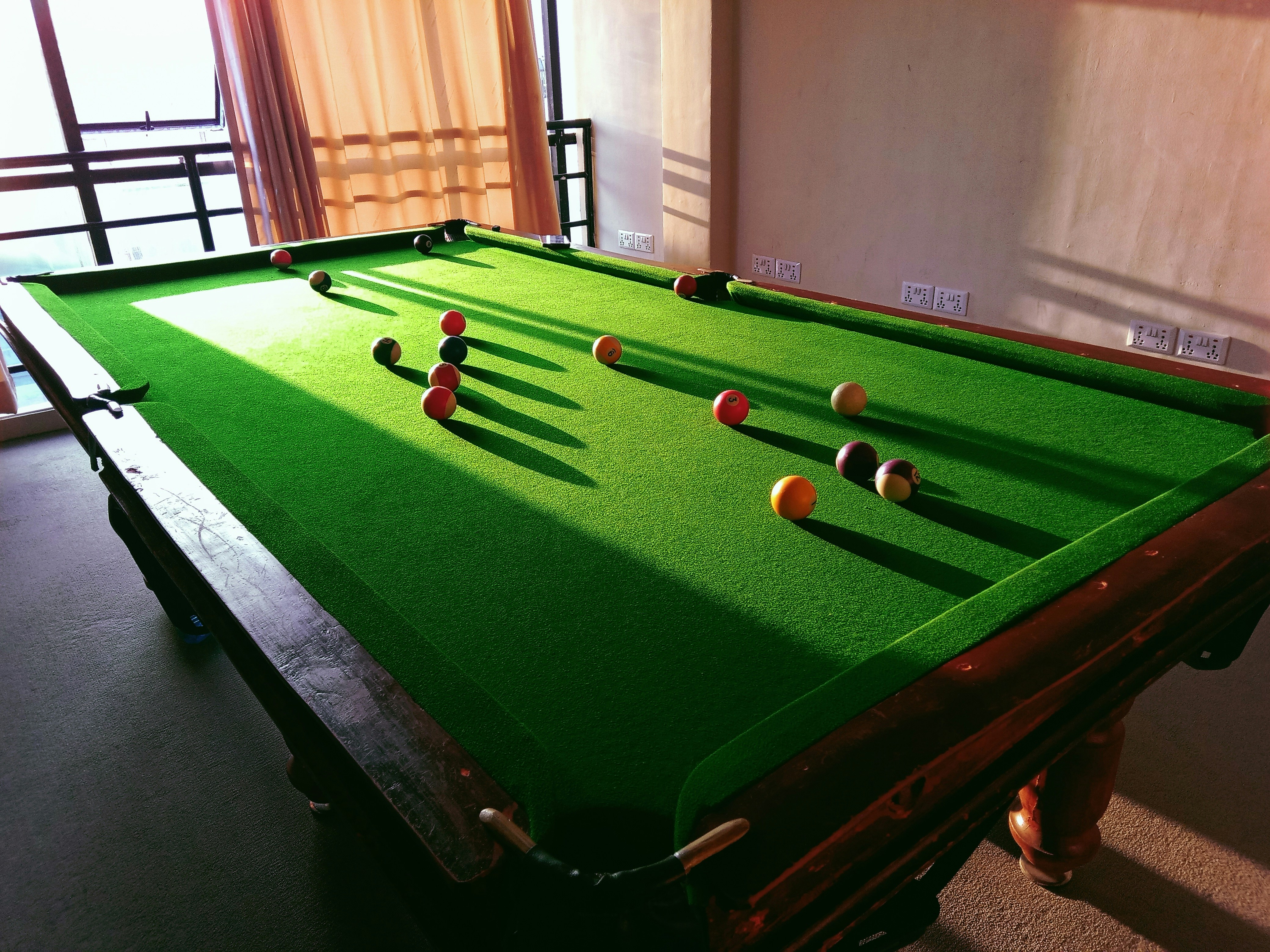 This picture shows a billiard table bathed in natural light coming through a window with orange curtains. The green felt of the table dramatically contrasts with the wood edges and the variably colored billiard balls strewn across it. Linear patterns of shadows from the panes of the window further animate the stillness of the surface. That is an interesting picture, as it merges indoor fun with natural light; thus, creating such a warm and appealing view that soothes the aesthetic sense just by looking at this particular leisure activity.