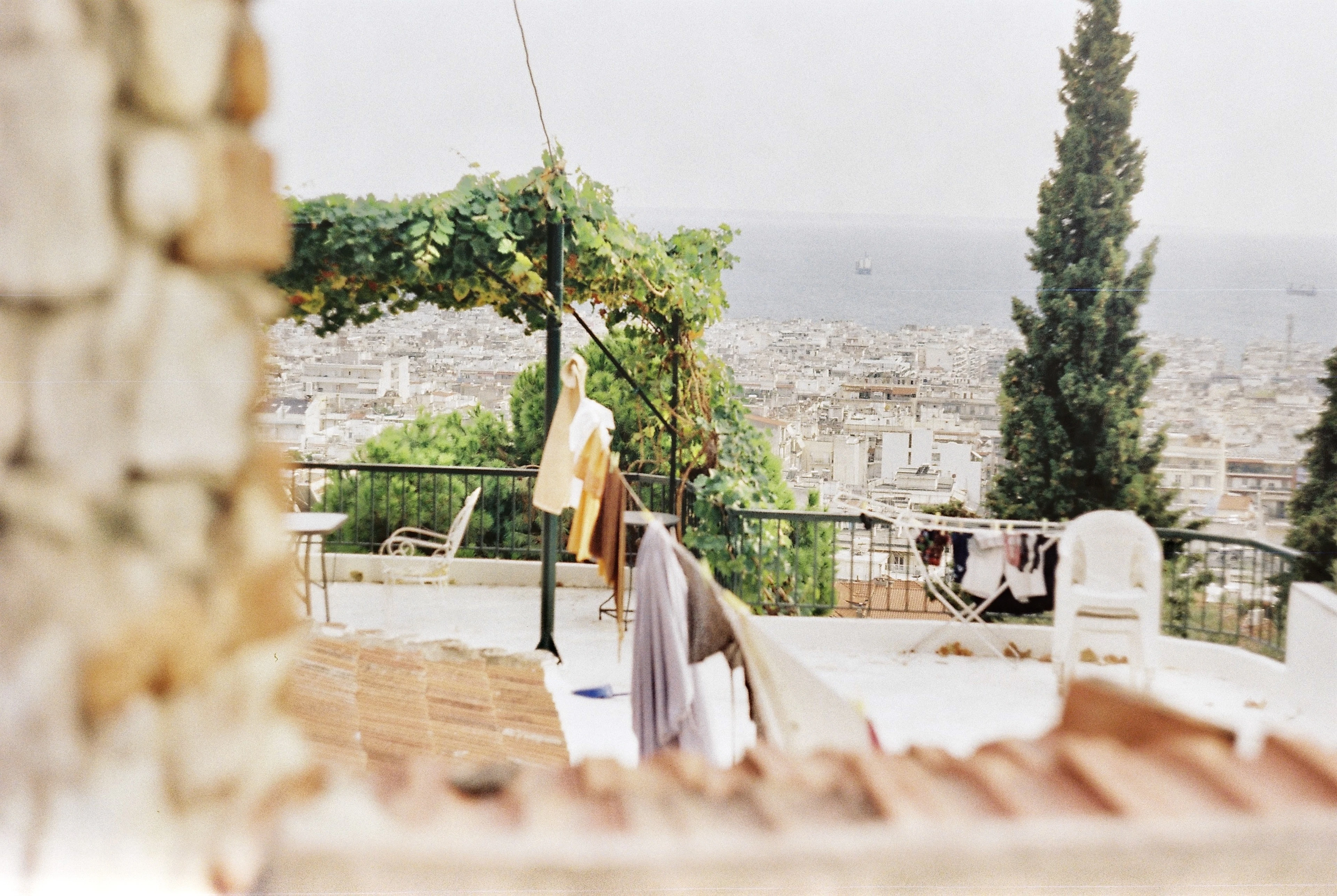 A whitewashed rooftop terrace in Thessaloniki's Upper Town, laundry and a vine-covered pergola in the foreground and the white city falling away to the Thermaic Gulf beyond.