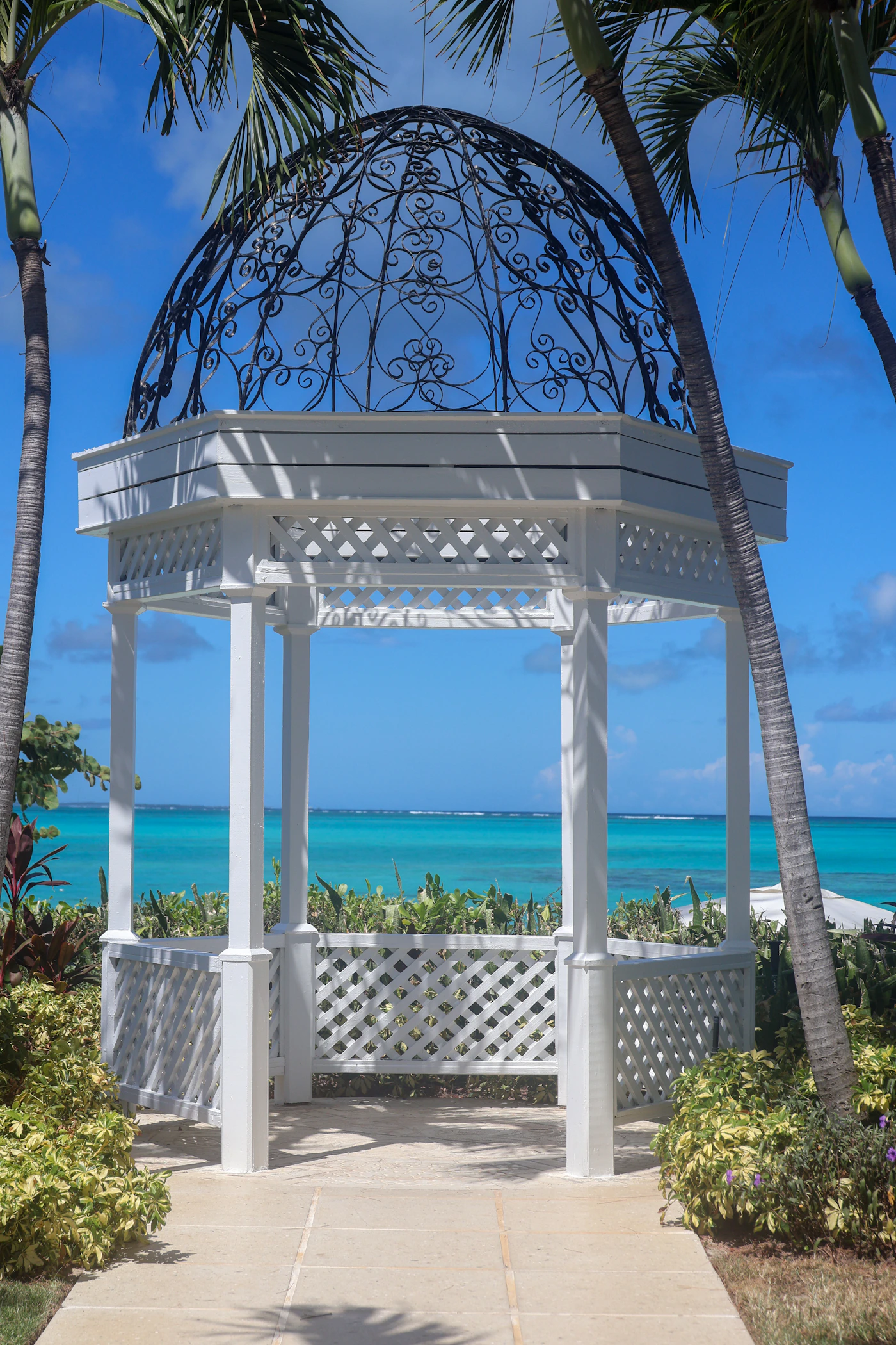 A white ornate wedding ceremony gazebo standing among palm trees with bright turquoise ocean visible behind it — the open-air ceremony structure that defines beachfront destination wedding venues