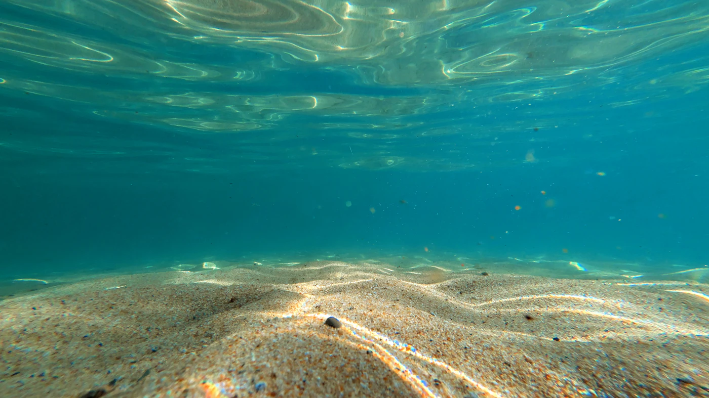 Fondo de arena bajo aguas poco profundas, con la luz del sol ondulando en la superficie
