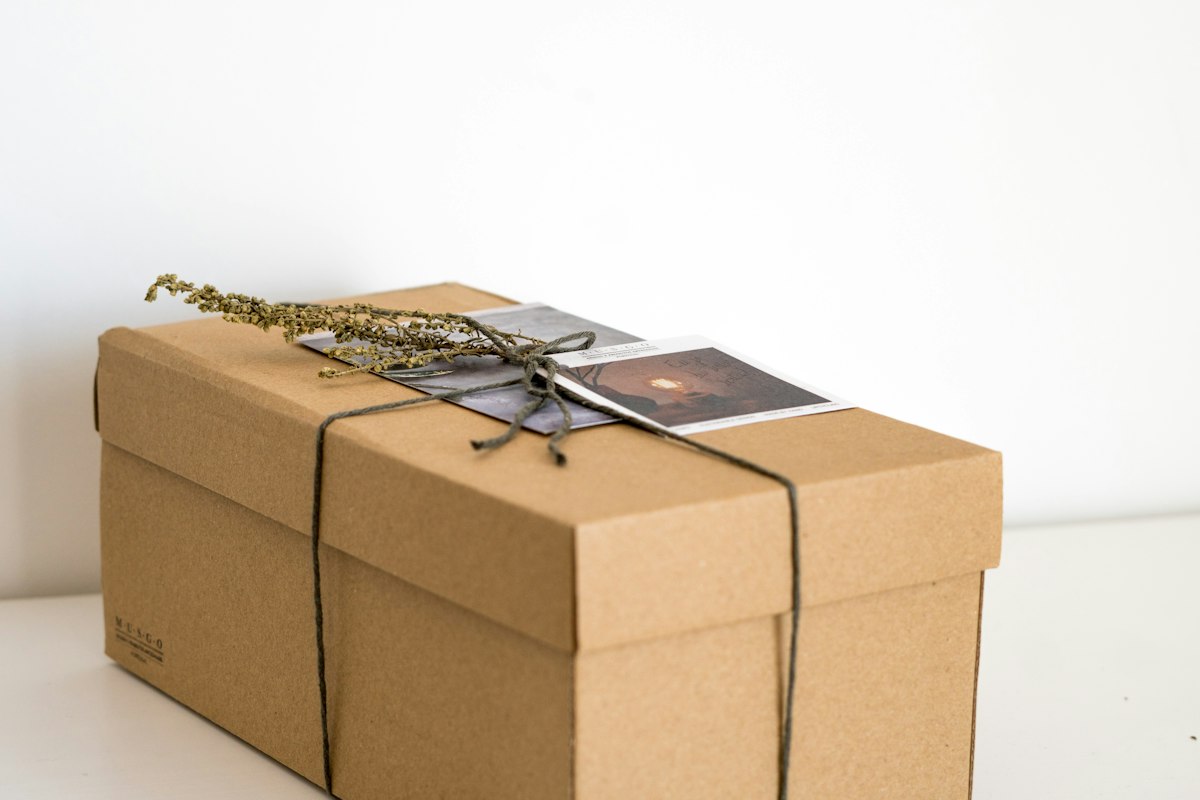 Boîte en carton kraft fermée par une ficelle, avec une carte et un brin de fleurs séchées posés sur le dessus, sur un meuble blanc