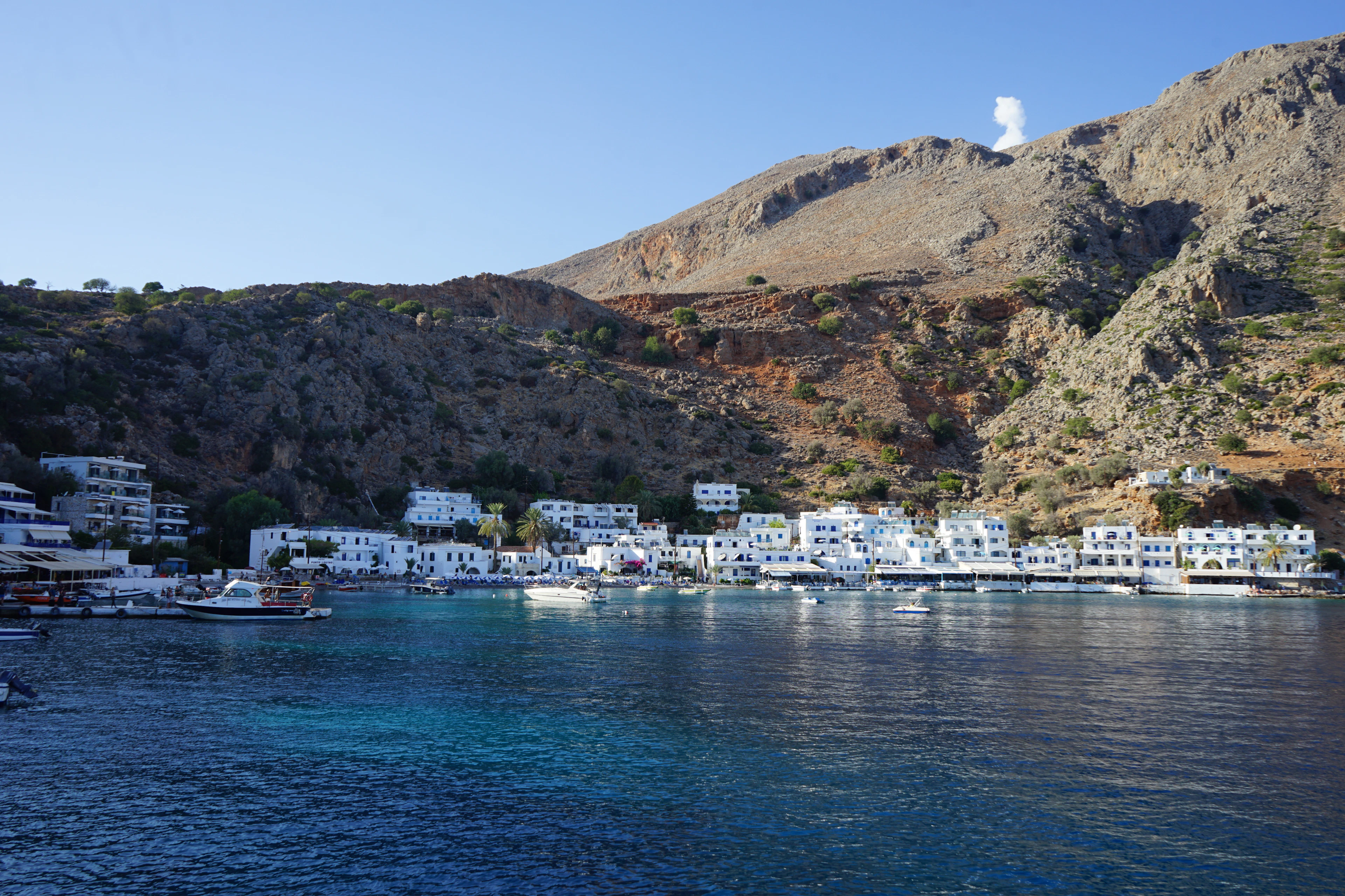 Whitewashed houses and a harbor cluster at the foot of a mountain in the seaside village of Loutro, Crete.