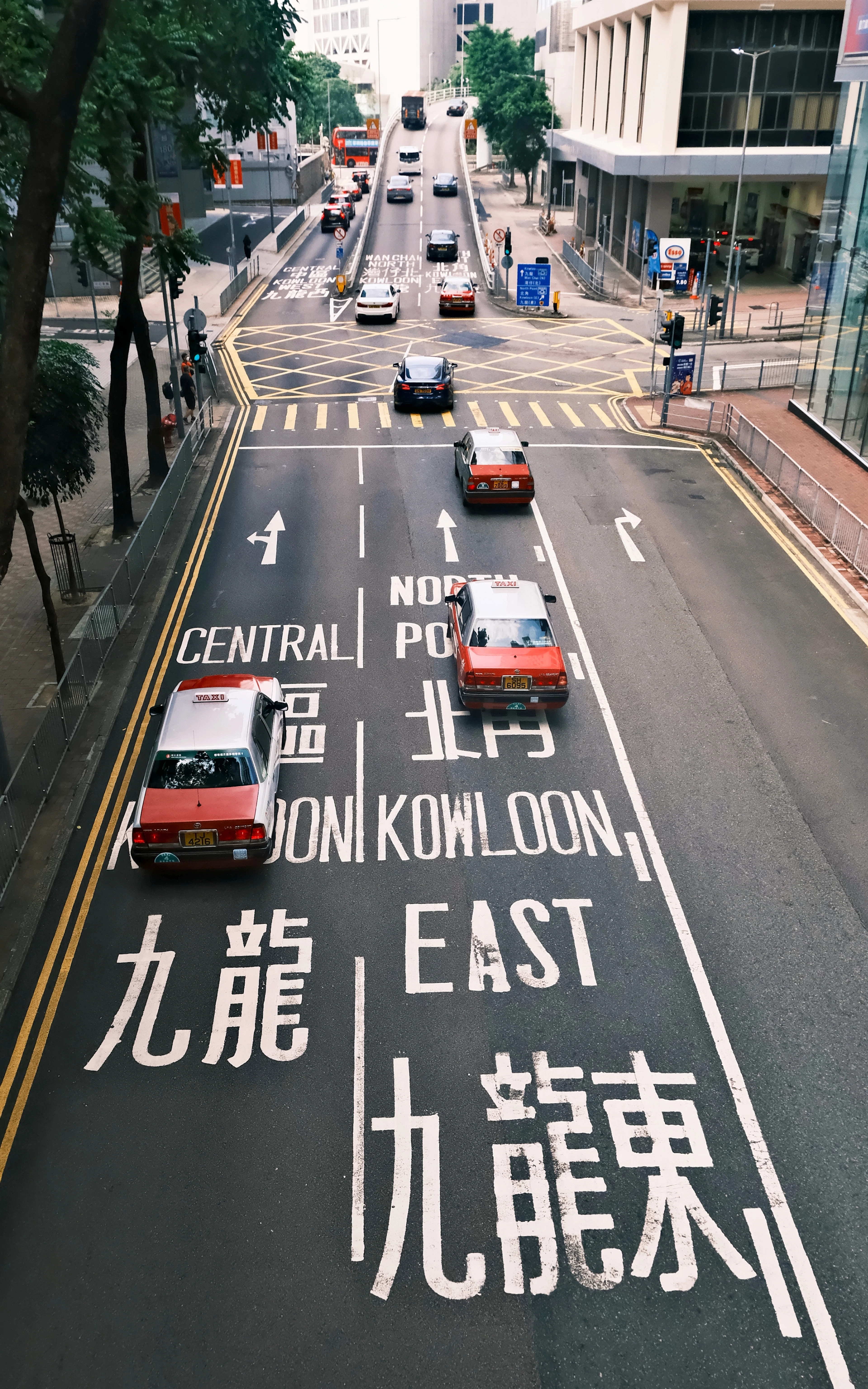 Red taxis cross a Hong Kong street painted with bilingual Central and Kowloon East lane markings.