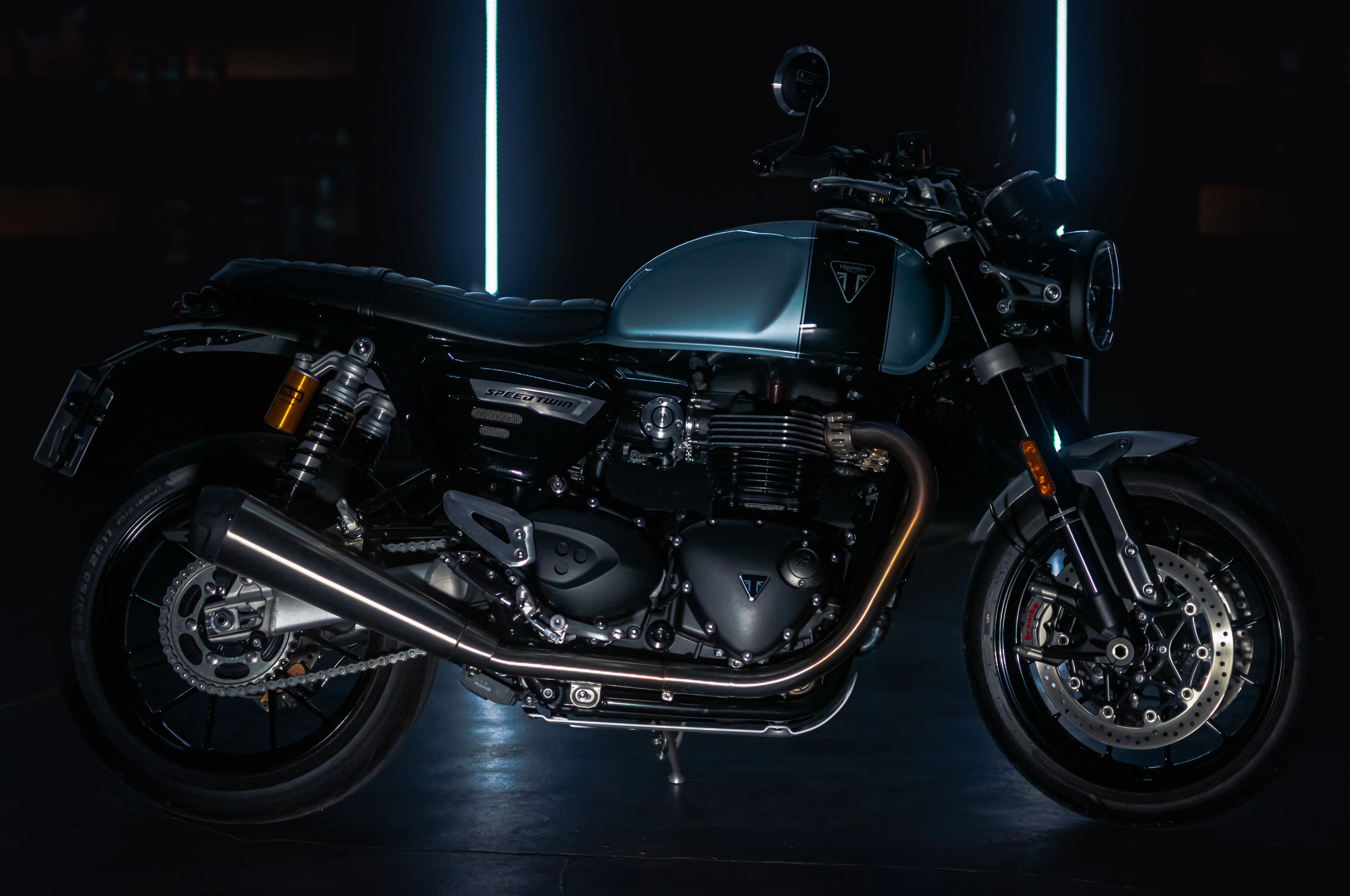 A black and dark blue Triumph Speed Twin motorcycle in a moody studio with vertical neon light strips, its "Speed Twin" badge visible on the side panel