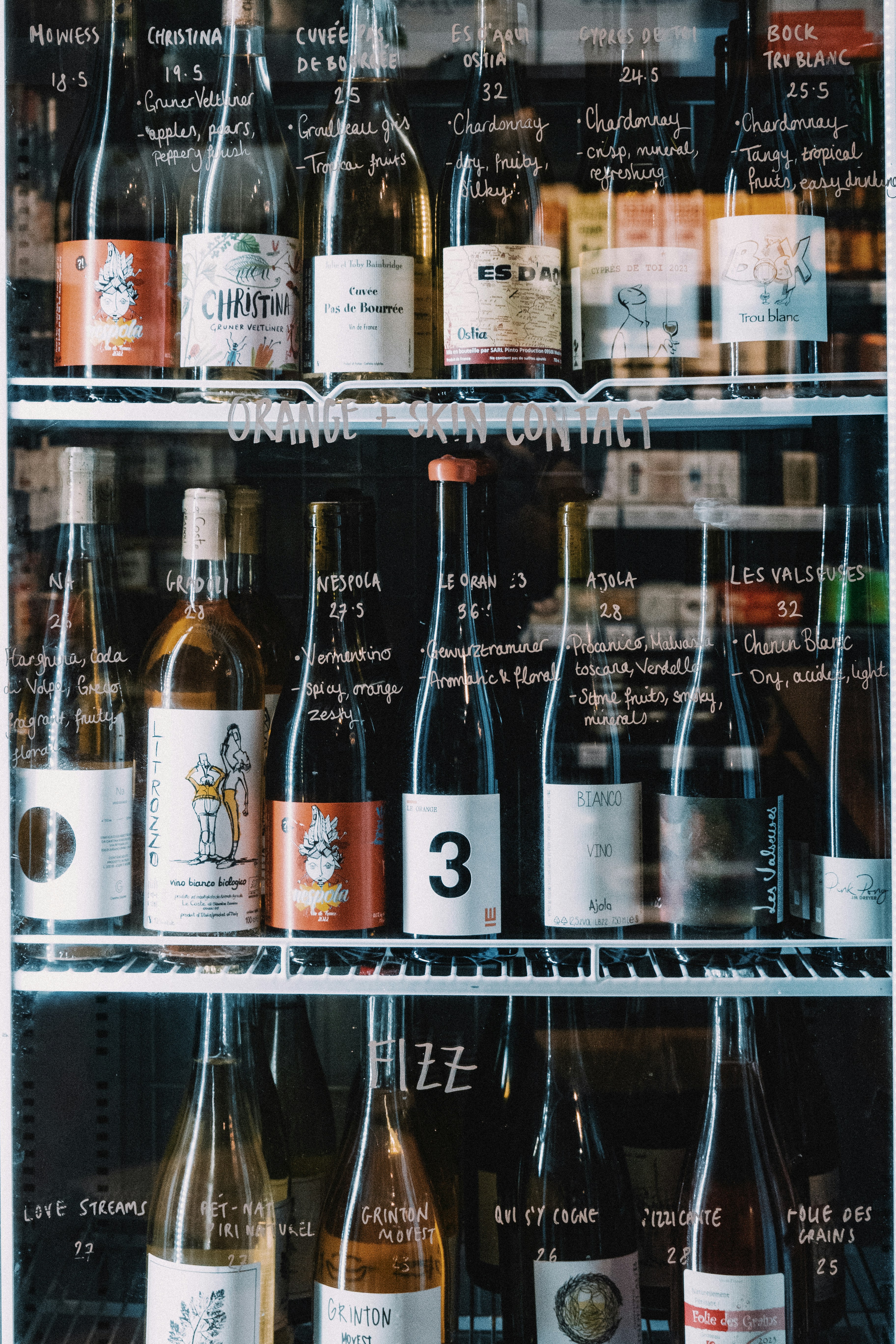 A row of wine bottles with handwritten chalkboard labels on a glass display, centered by a bottle bearing a bold '3'.