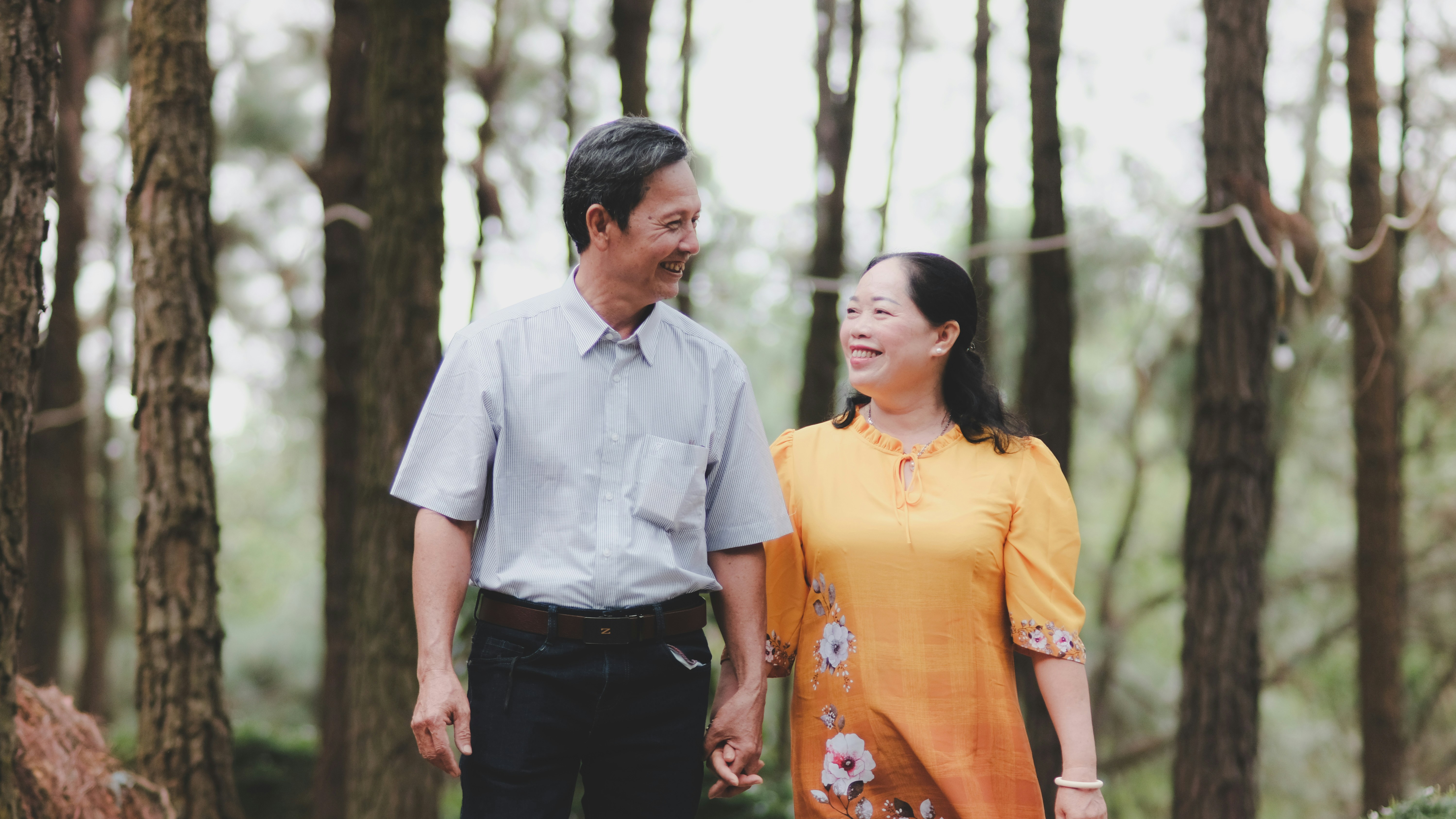Happy couple in their 50s walking together on a bright tree-lined path, warm afternoon light, genuine laughter and connection