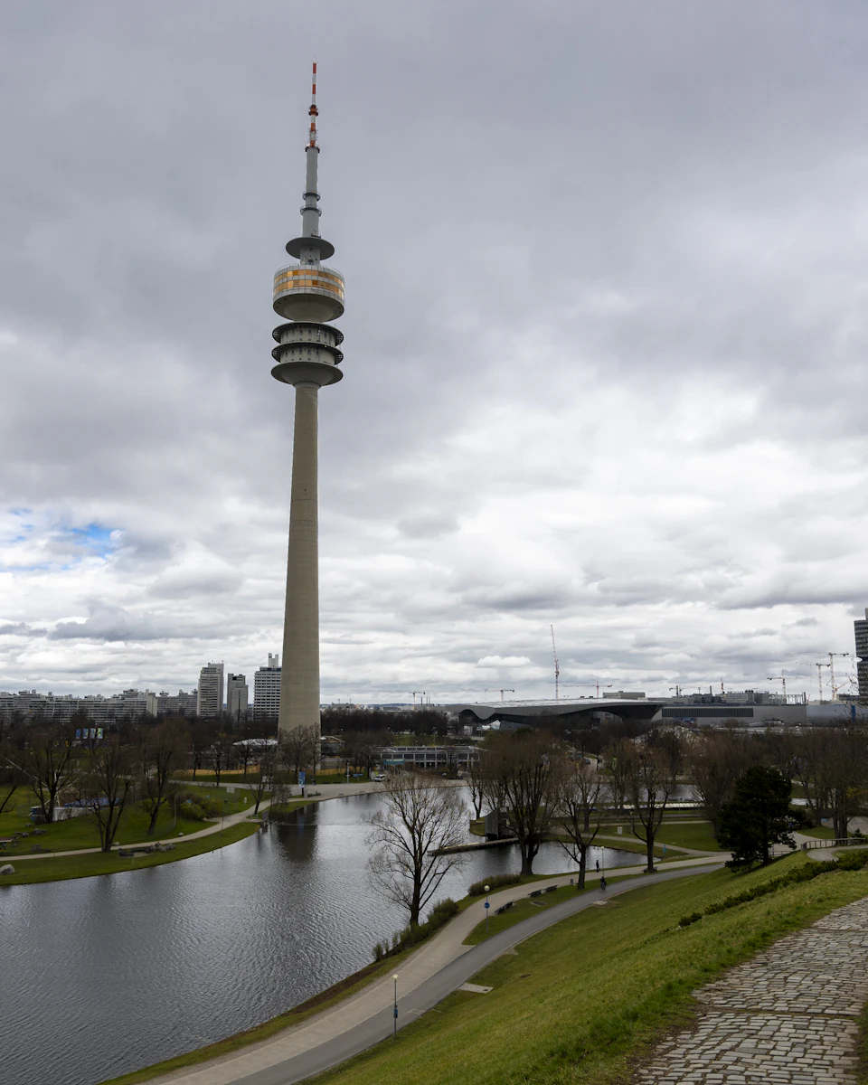 The image shows a tall, gray tower with a red and white antenna on top, standing against an overcast sky. The tower is situated next to a large body of water, which reflects the surrounding cityscape. In the foreground, there are trees lining the path leading up to the tower, creating a sense of depth in the scene.

The image also contains text that reads "sa_1896".