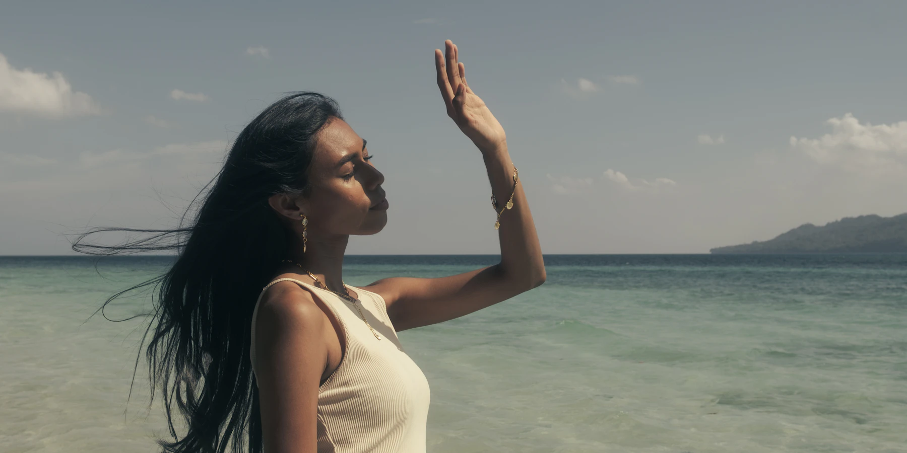 Adult woman at the ocean's edge in golden light wearing gold earrings, a fine necklace and bracelet