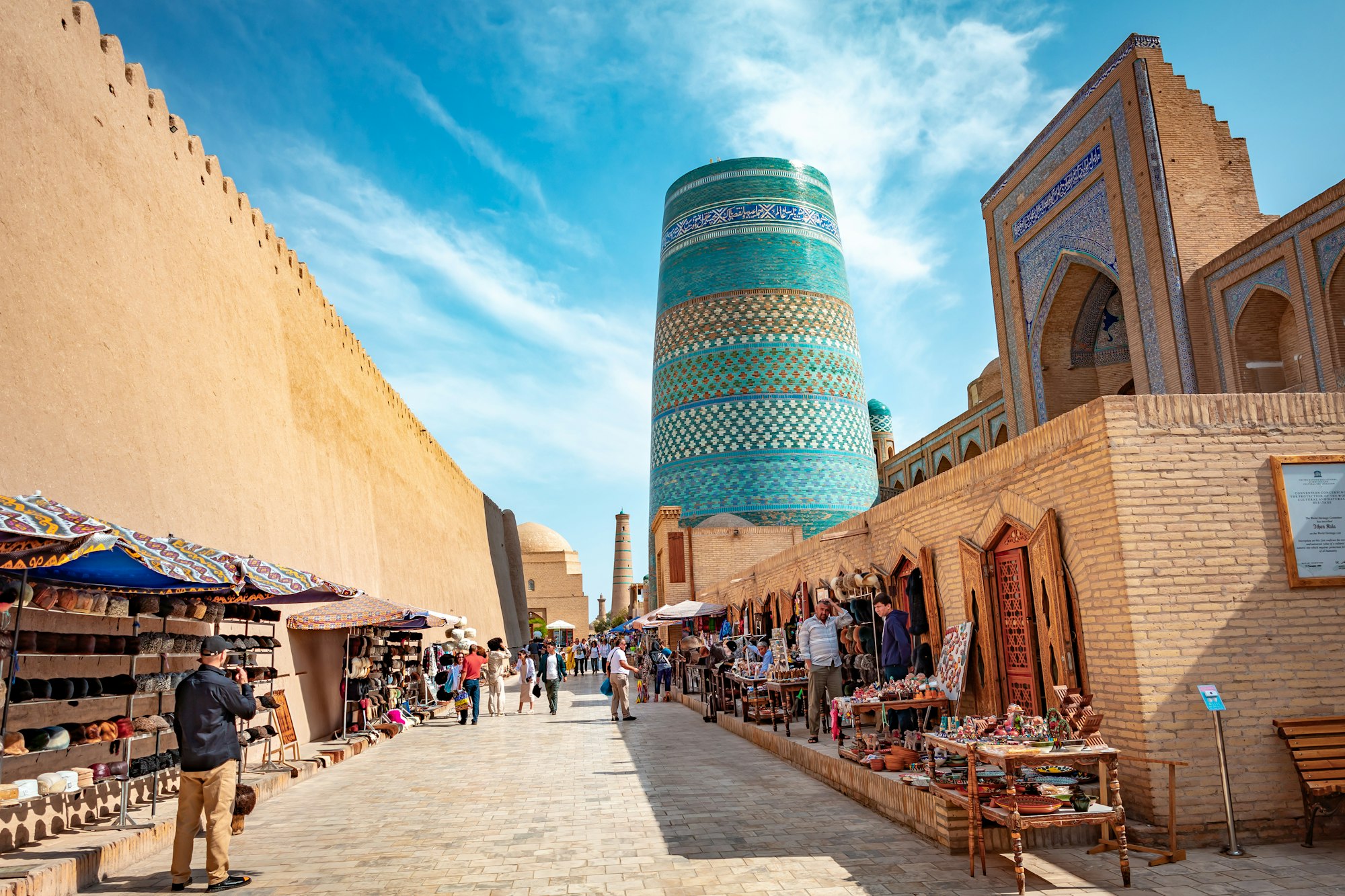 مناره‌ی کوتاه و کاشی‌کاری‌شده‌ی کلته‌منار در انتهای کوچه‌ای با بساط صنایع دستی در خیوه