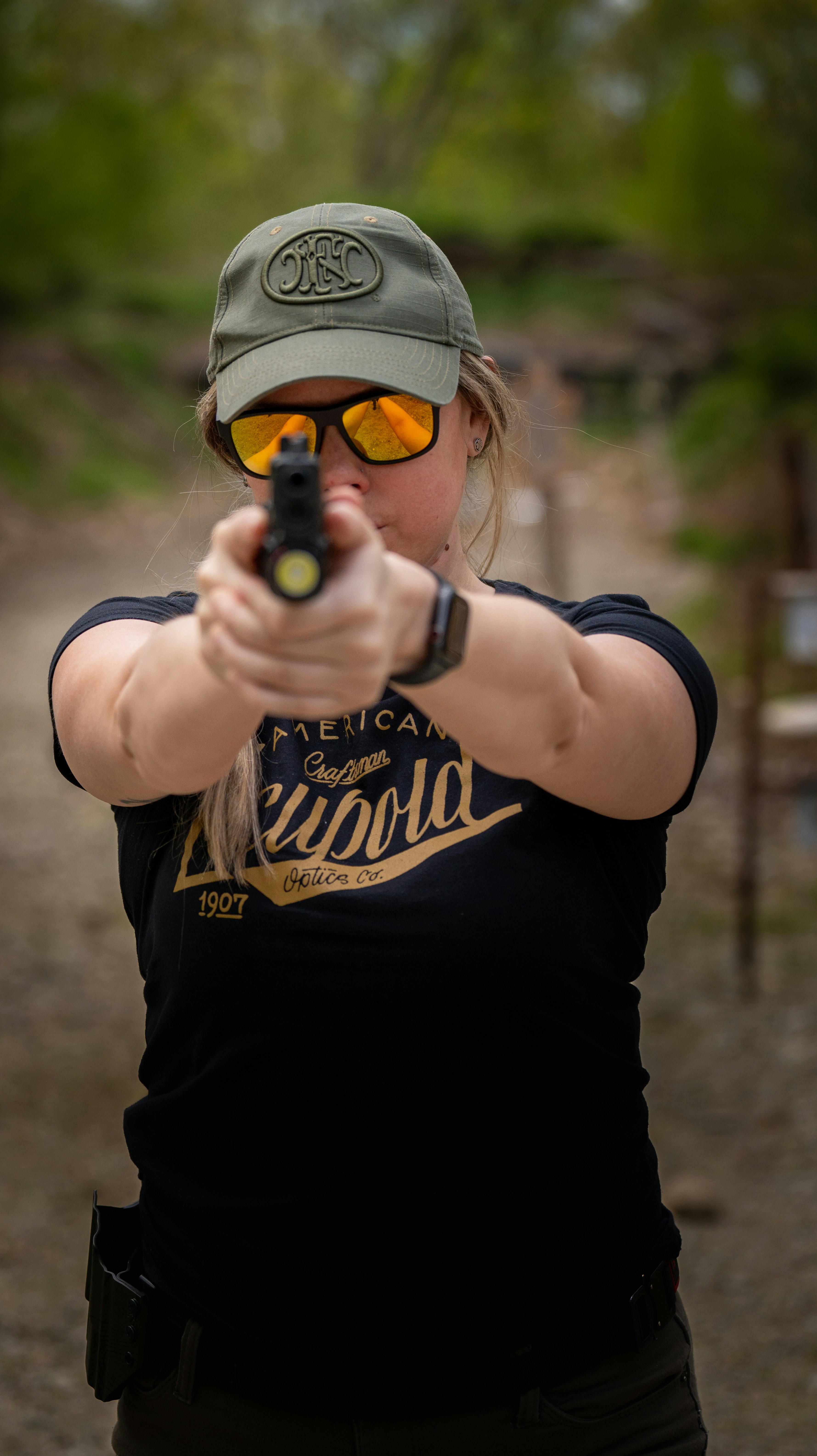 A woman with a hat and sunglasses aiming a gun