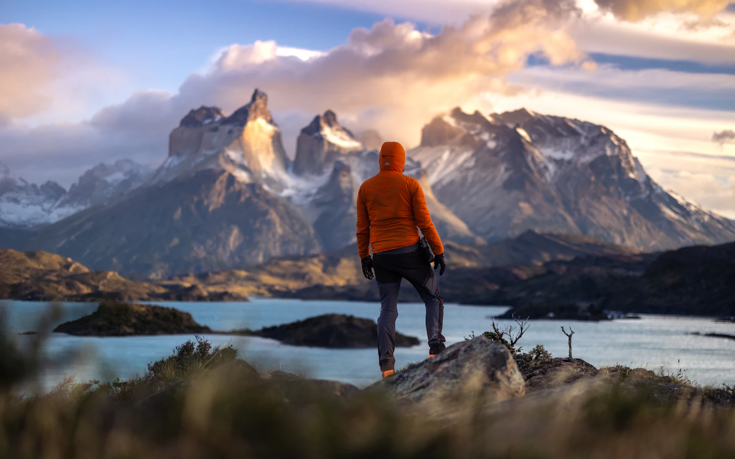 A hiker in an orange jacket standing on a ridge facing the granite towers of Torres del Paine.